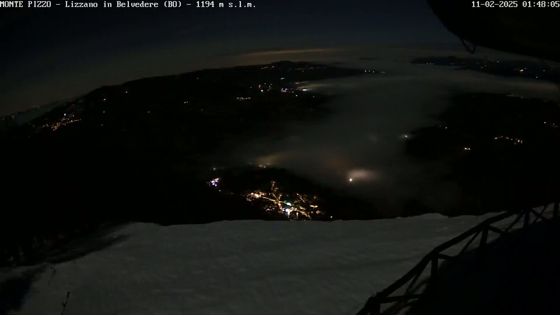 A nighttime, snowy, high-altitude view of a valley shrouded in fog, with the lights of a town visible below.