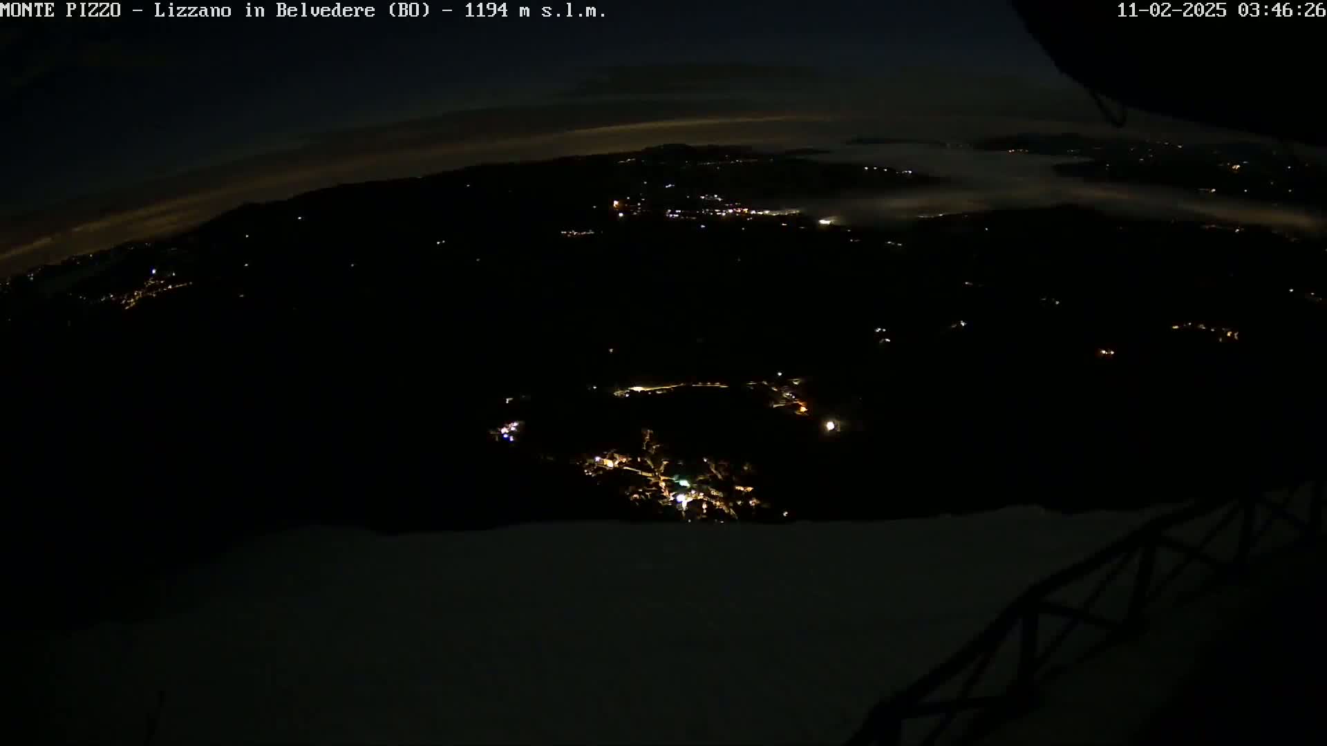 A nighttime, high-angle view shows a dark landscape with sparsely scattered lights below, and a layer of fog or clouds in the distance.