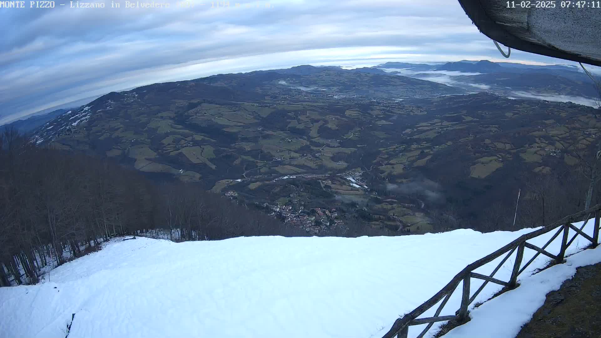 A snowy hillside overlooks a valley with a town nestled among rolling hills and low-lying fog under a cloudy sky.