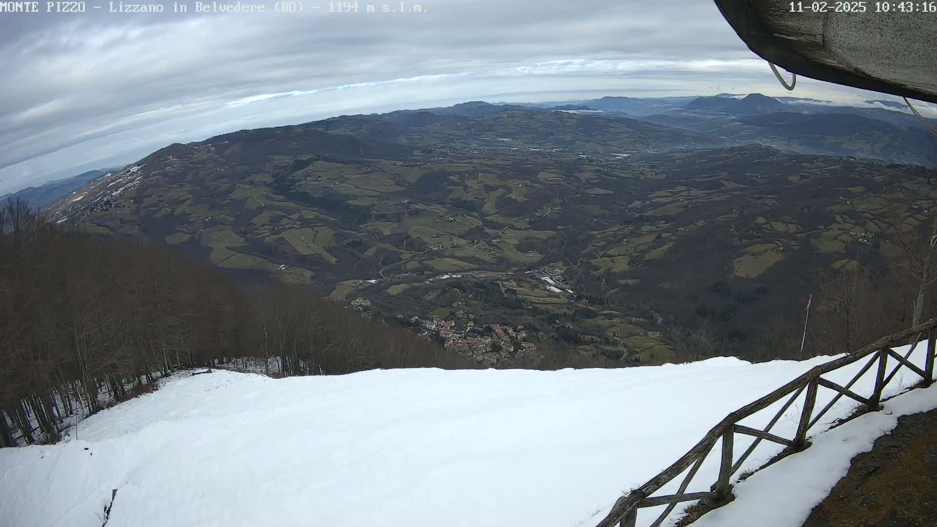 A snowy mountain slope overlooks a valley of dark green and brown hills under a mostly cloudy sky.