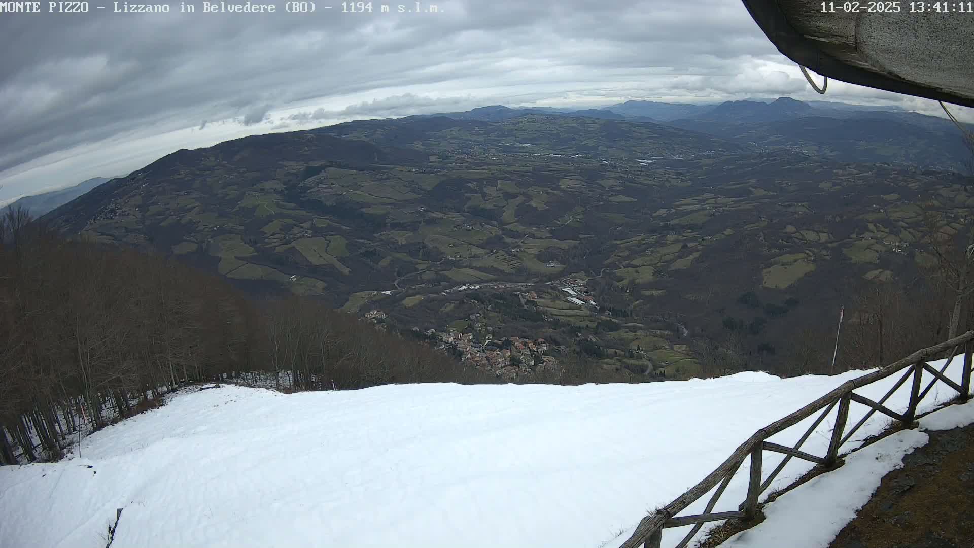 A snow-covered ski slope overlooks a valley of dark green hills and a small town under a cloudy sky.