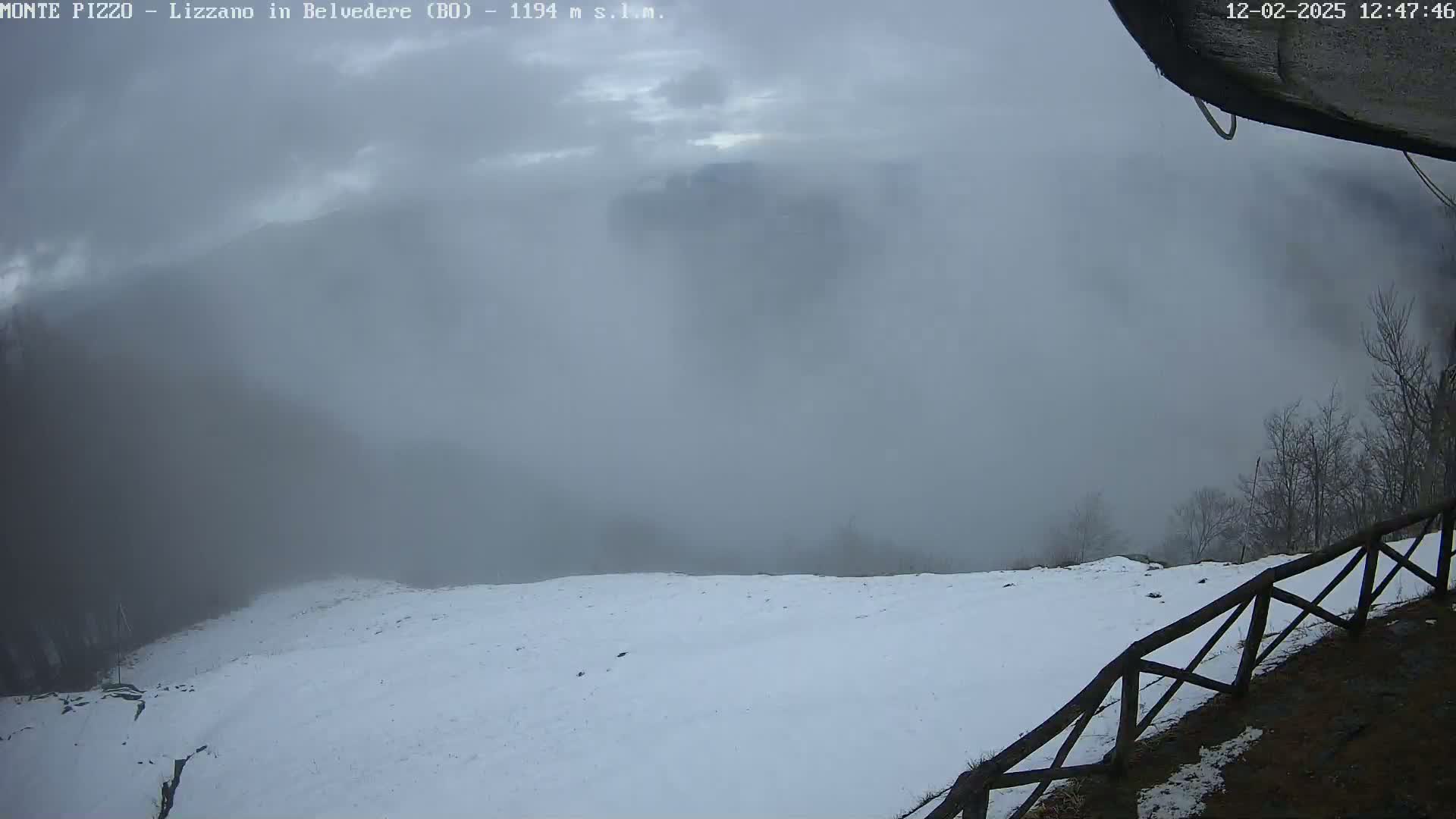 A snowy landscape is shrouded in fog, with a wooden fence visible in the foreground.