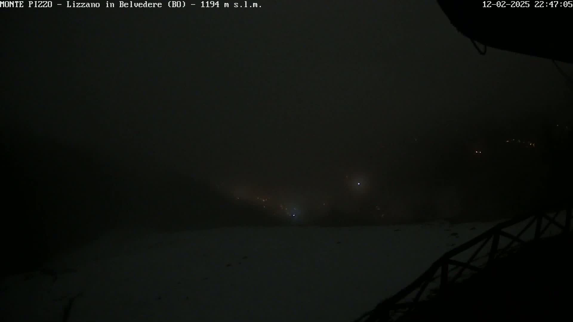 A nighttime, foggy view shows a snowy landscape with a few distant lights visible through the haze.