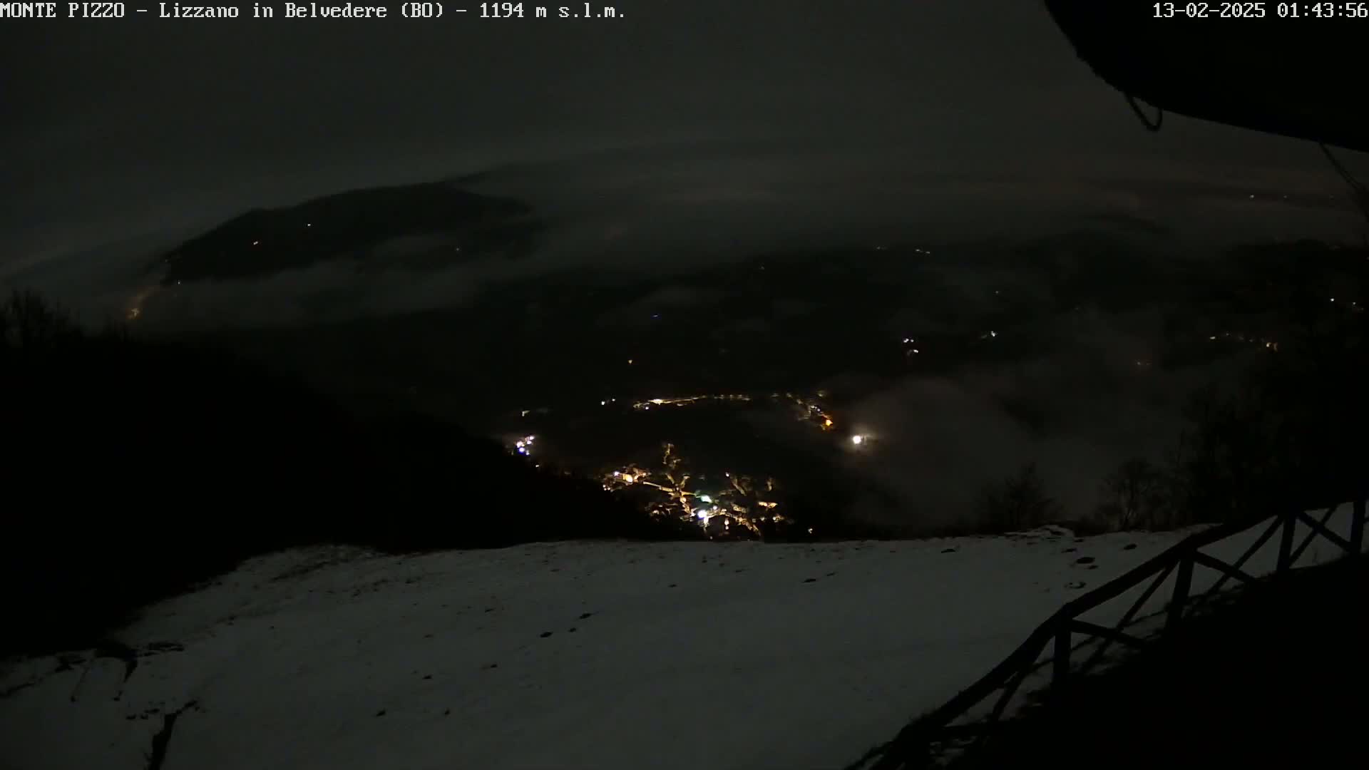 A nighttime view of a snow-covered landscape shows a valley with distant lights below, partially obscured by fog or low clouds.
