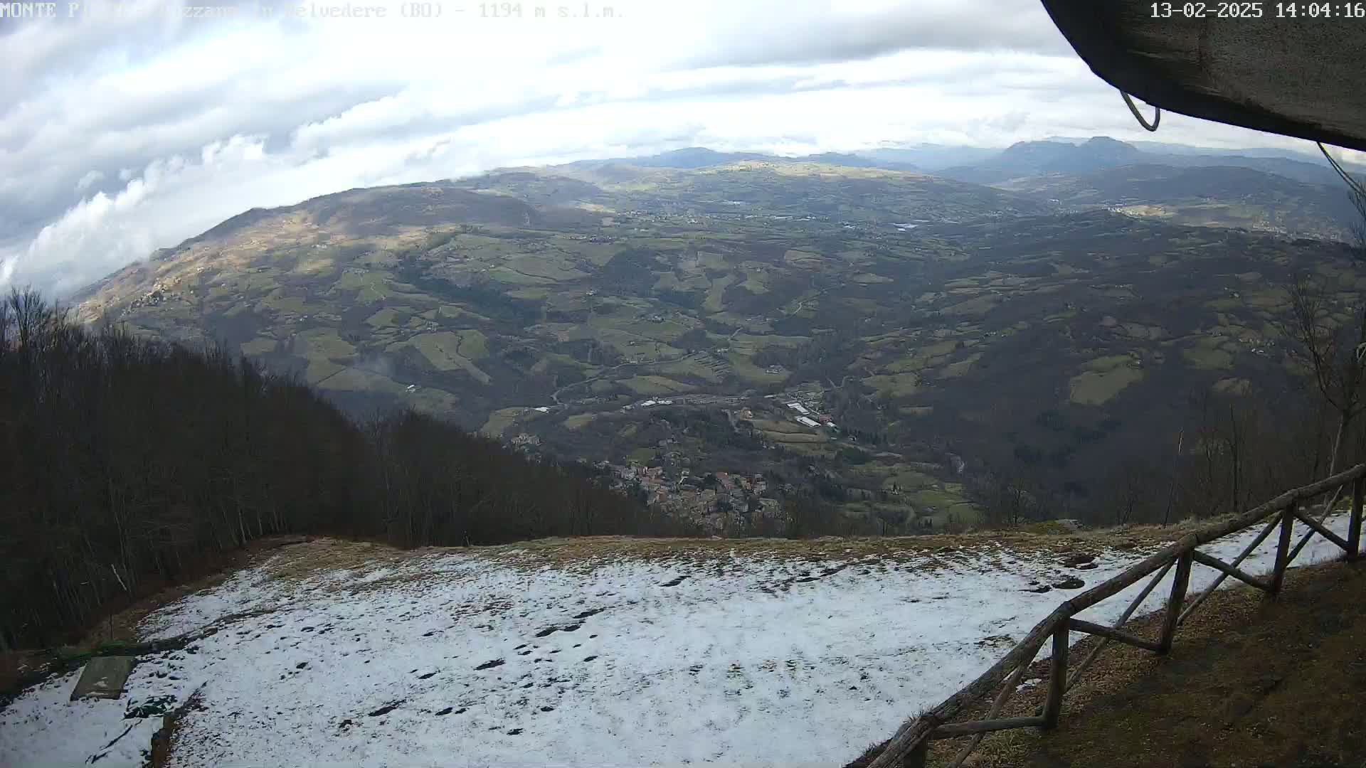 A snowy, elevated viewpoint overlooks a vast valley of rolling hills and a small village under a cloudy sky.