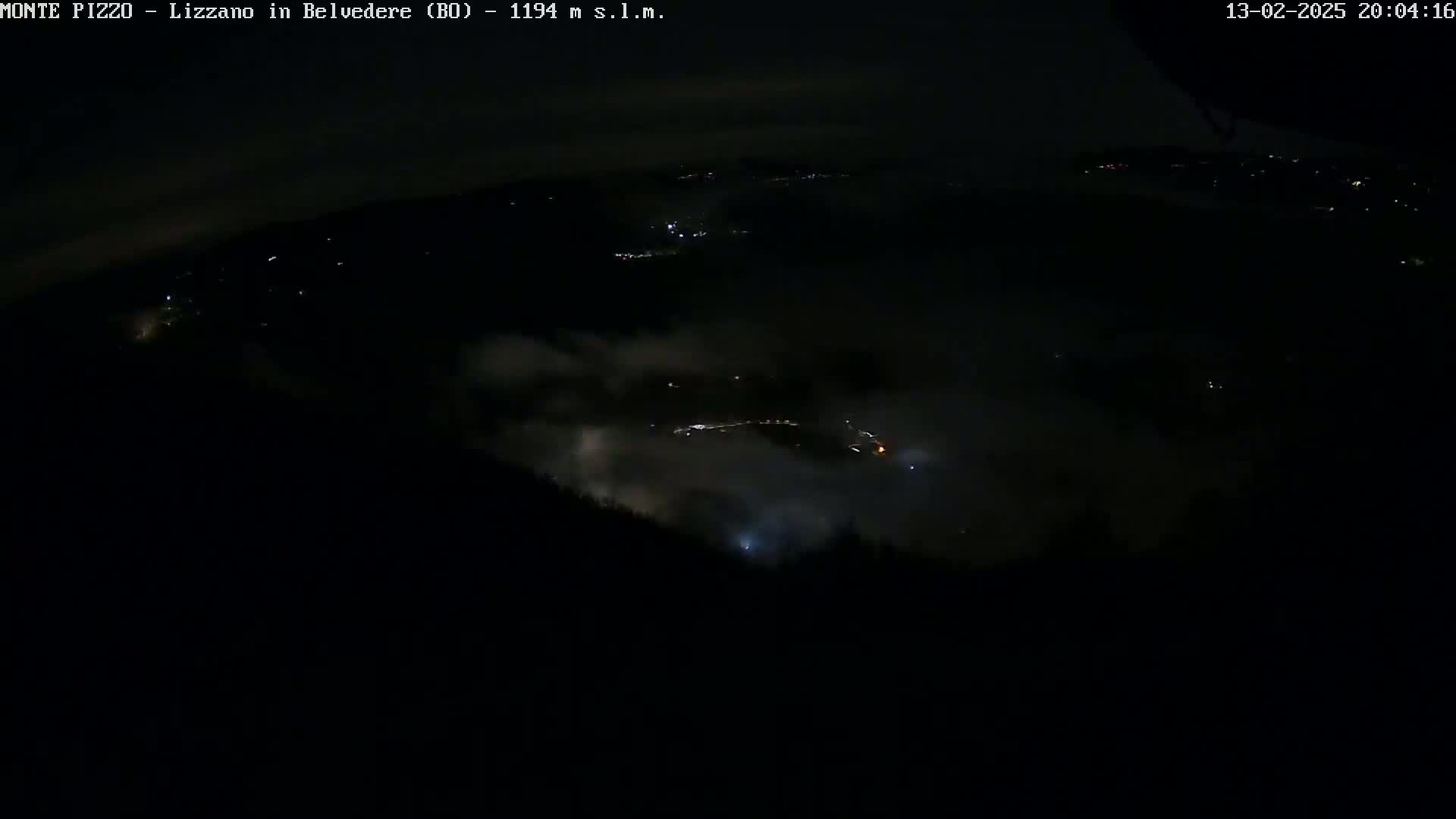 A nighttime, high-angle view shows a low-lying layer of fog or clouds obscuring the lower areas, with scattered lights visible in the distance and a dark foreground of trees or hills.