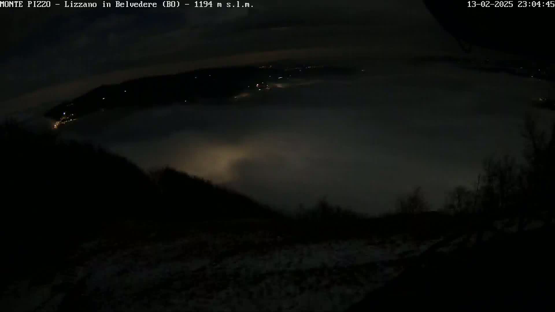 A nighttime, low-visibility view of a valley covered in fog, with a few lights visible in the distance and some snow on the ground in the foreground.