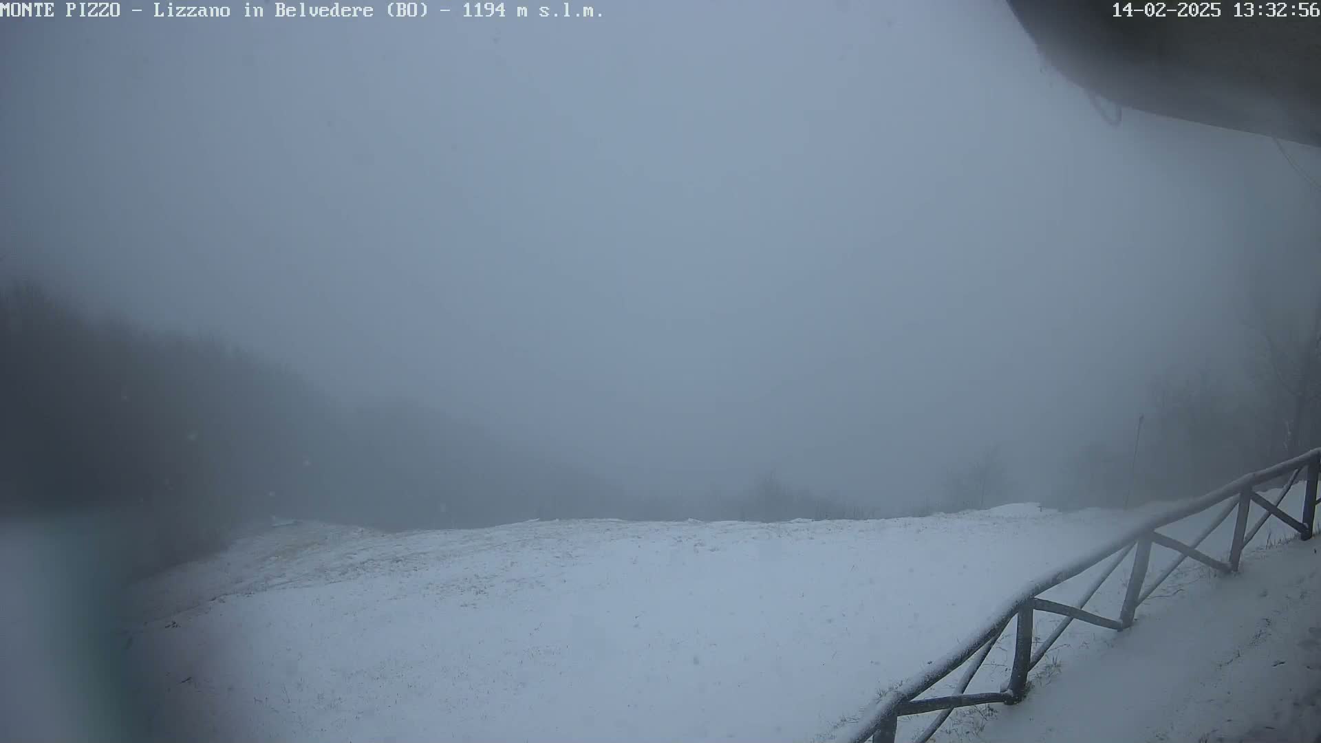 A snow-covered landscape is shrouded in heavy fog, with a wooden fence partially visible in the foreground.