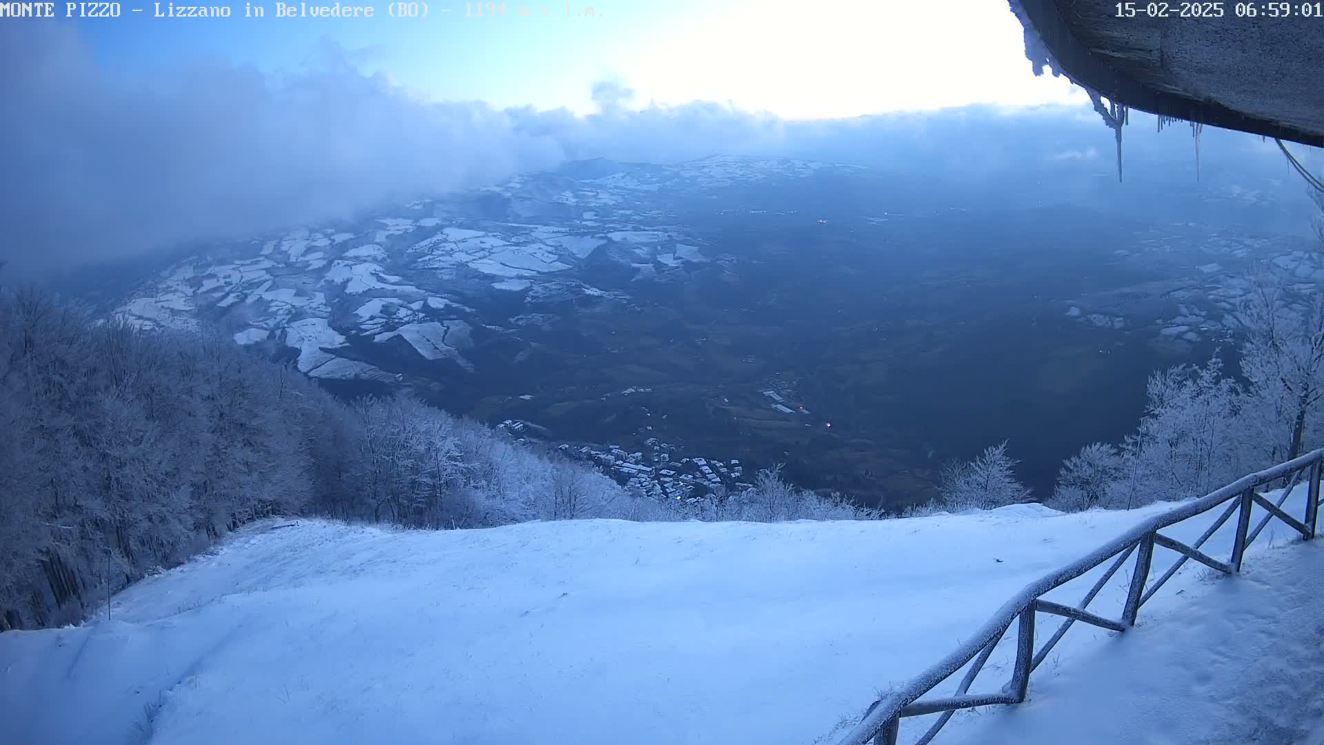 A snow-covered landscape viewed from a snowy mountaintop reveals a valley with patches of snow, a small town, and a wooden fence in the foreground.