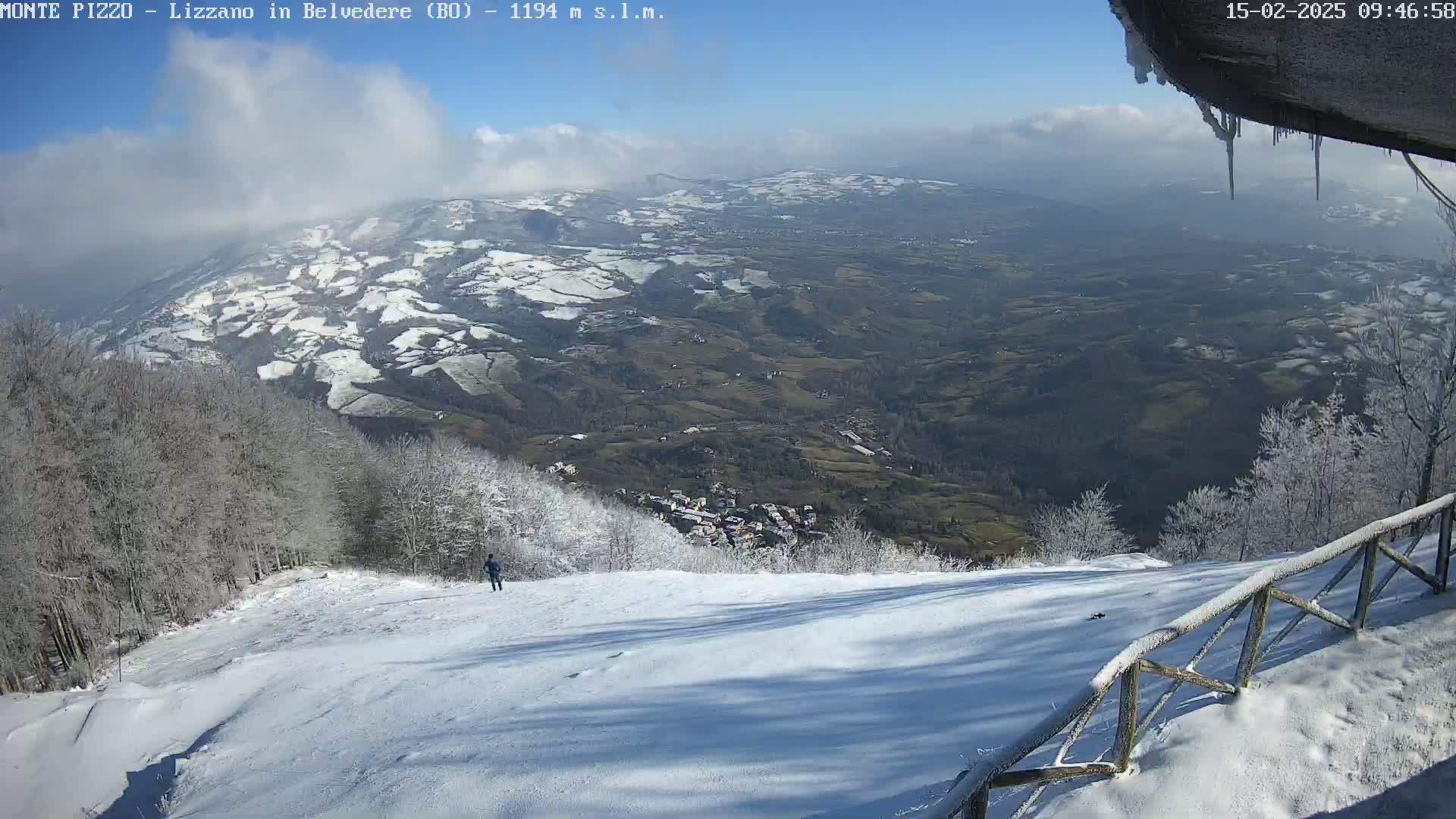 A snowy, mountainous landscape under a mostly clear blue sky is viewed from a high vantage point, showing a snow-covered slope with a wooden fence and a lone person in the distance.