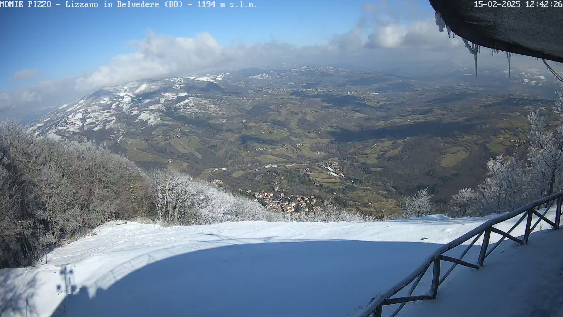 A snowy mountaintop view reveals a valley below dotted with buildings and farmland under a partly cloudy, sunny sky.