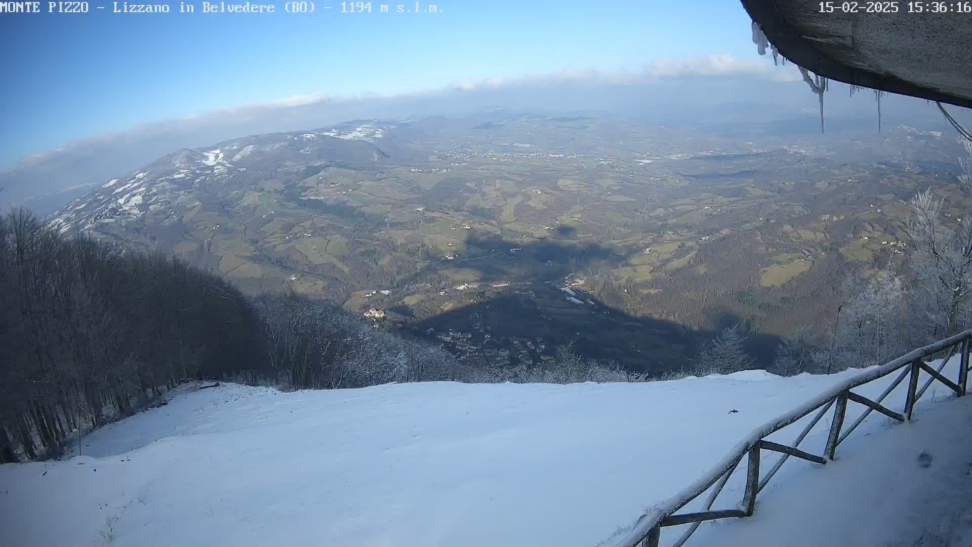 A snowy, mountainous landscape is viewed from a high vantage point on a partly sunny day, with a wooden fence in the foreground and icicles hanging from an overhanging structure.