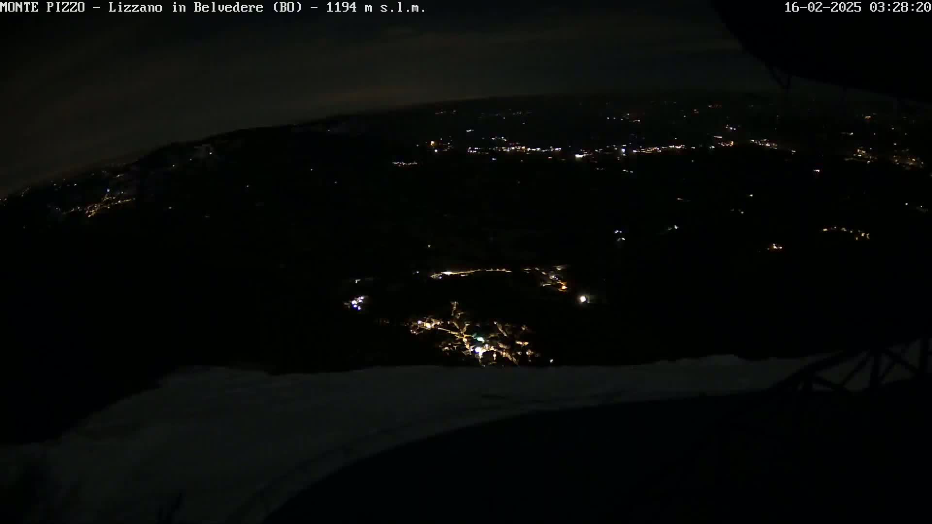 A nighttime, high-angle view shows a dark landscape with scattered lights from a town or village below, under a mostly clear, dark sky.