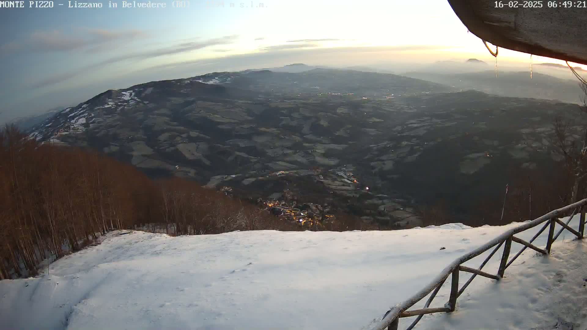 A snow-covered hillside overlooks a valley town at sunrise, with a wooden fence in the foreground.