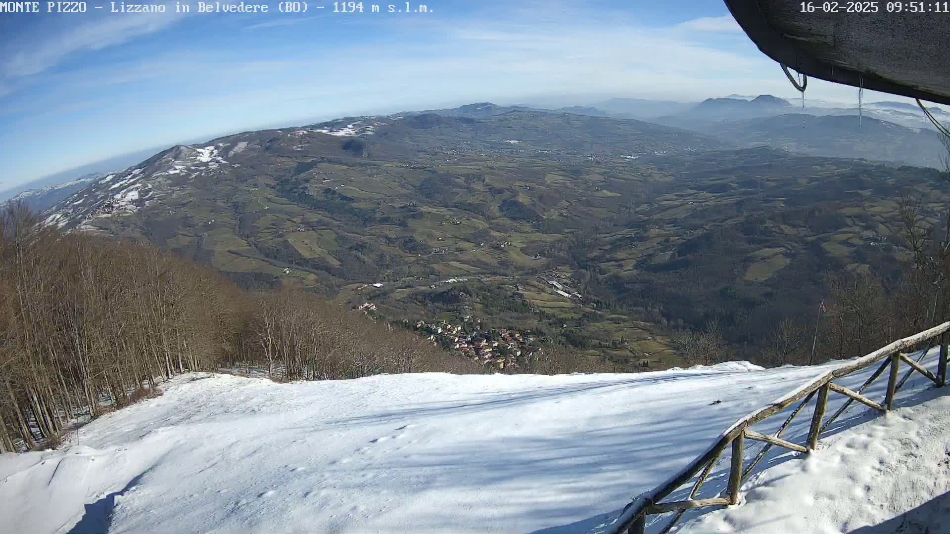 A snow-covered hillside overlooks a vast valley of farmland and a small village under a clear, sunny sky.