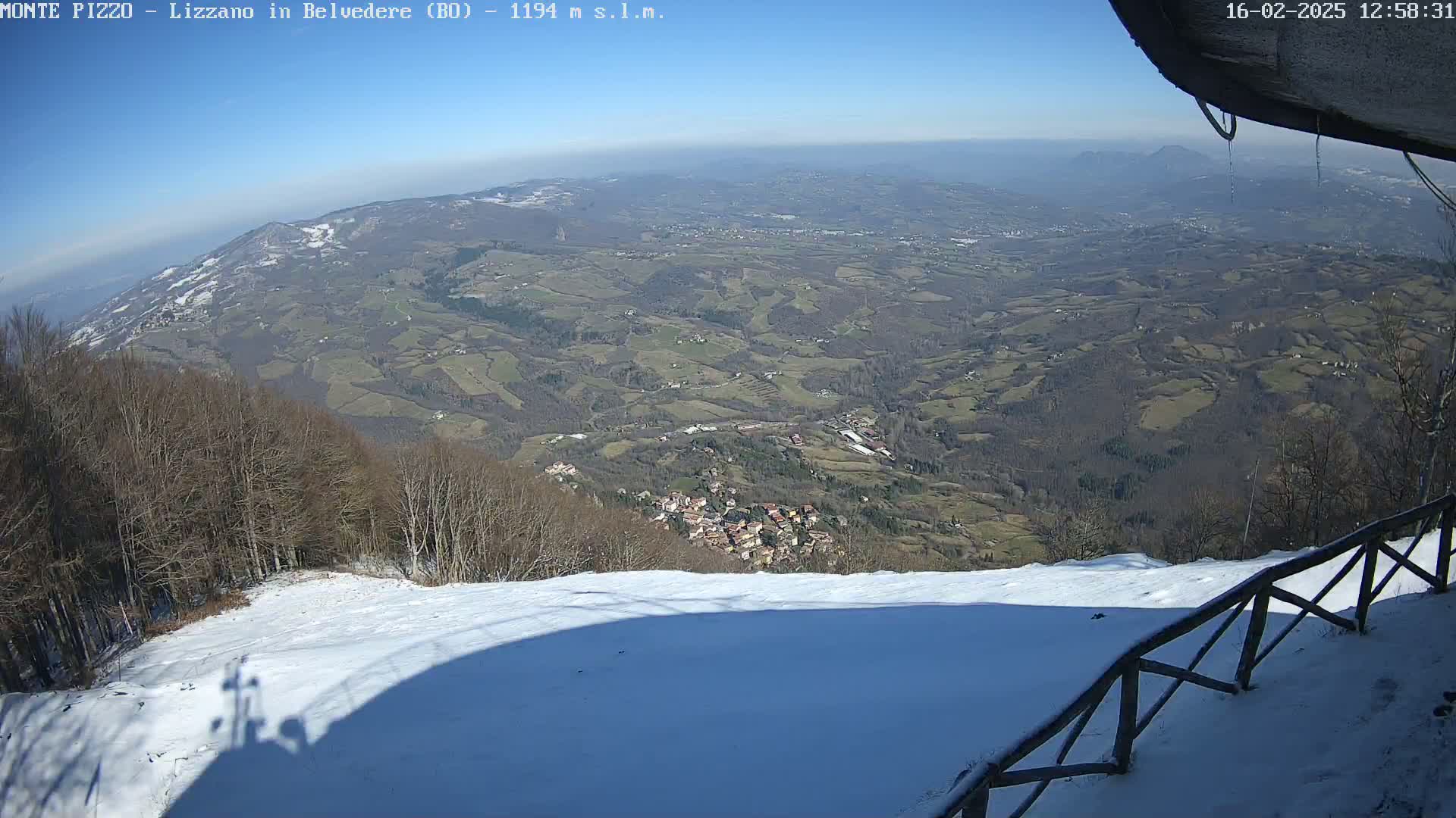 A snow-covered slope overlooks a vast valley of farmland and a small village under a clear blue sky.