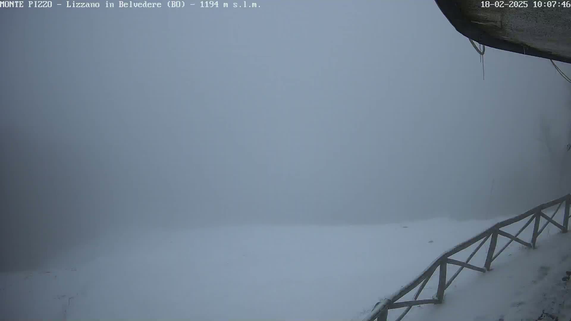 A snow-covered landscape is obscured by heavy fog, with a wooden fence partially visible in the lower right corner.
