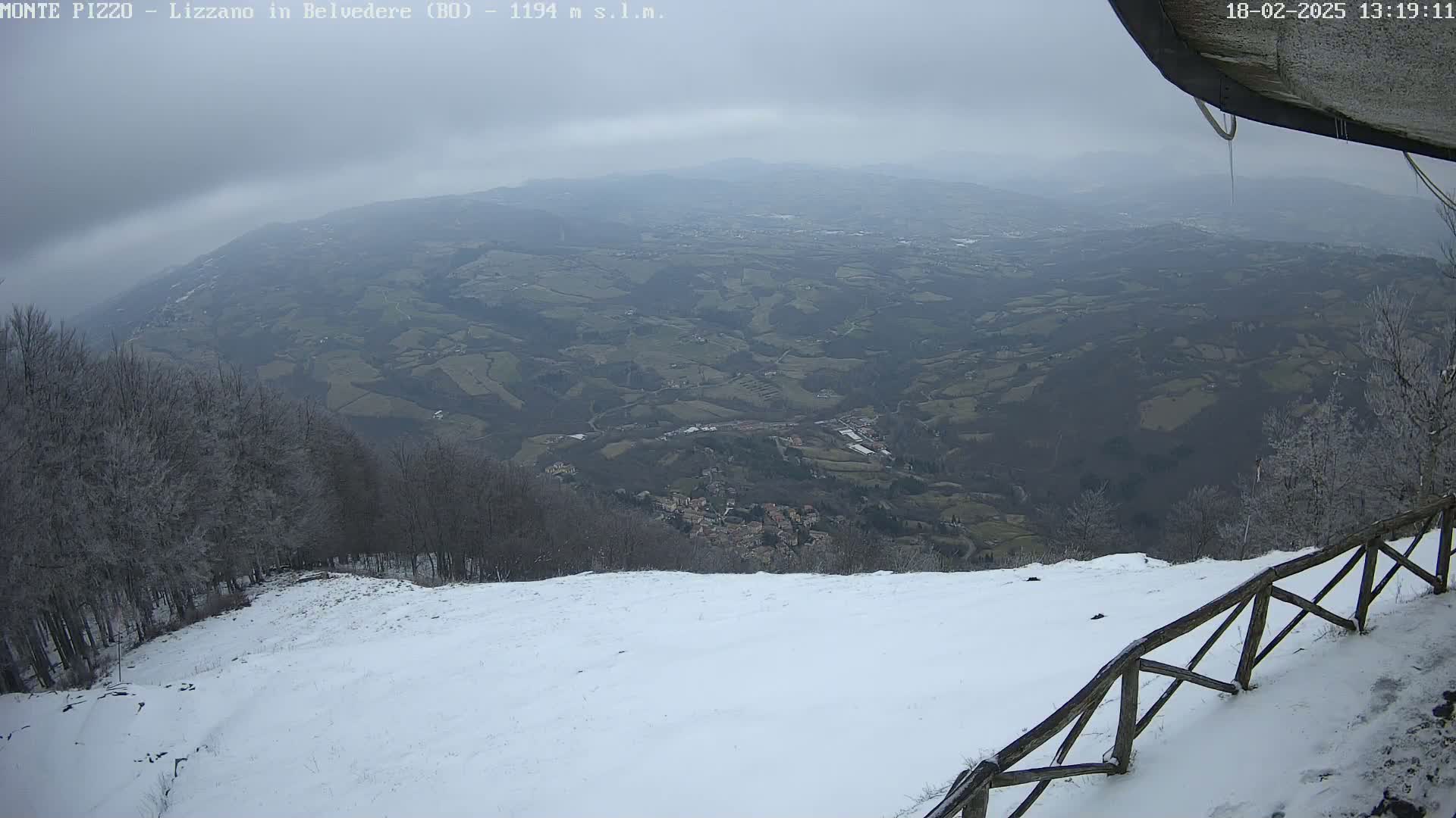 A snow-covered hillside overlooks a valley of farmland and a small town under an overcast sky.