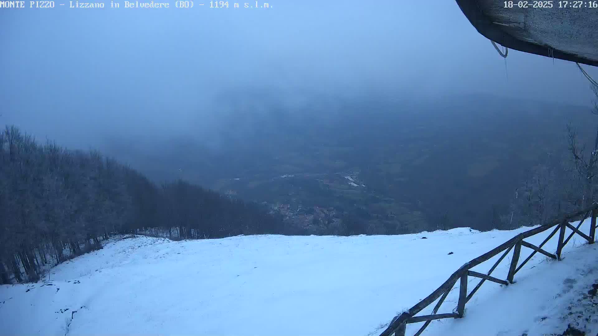 A snow-covered hillside slopes down to a hazy valley in the distance, with a wooden fence in the foreground under a foggy sky.