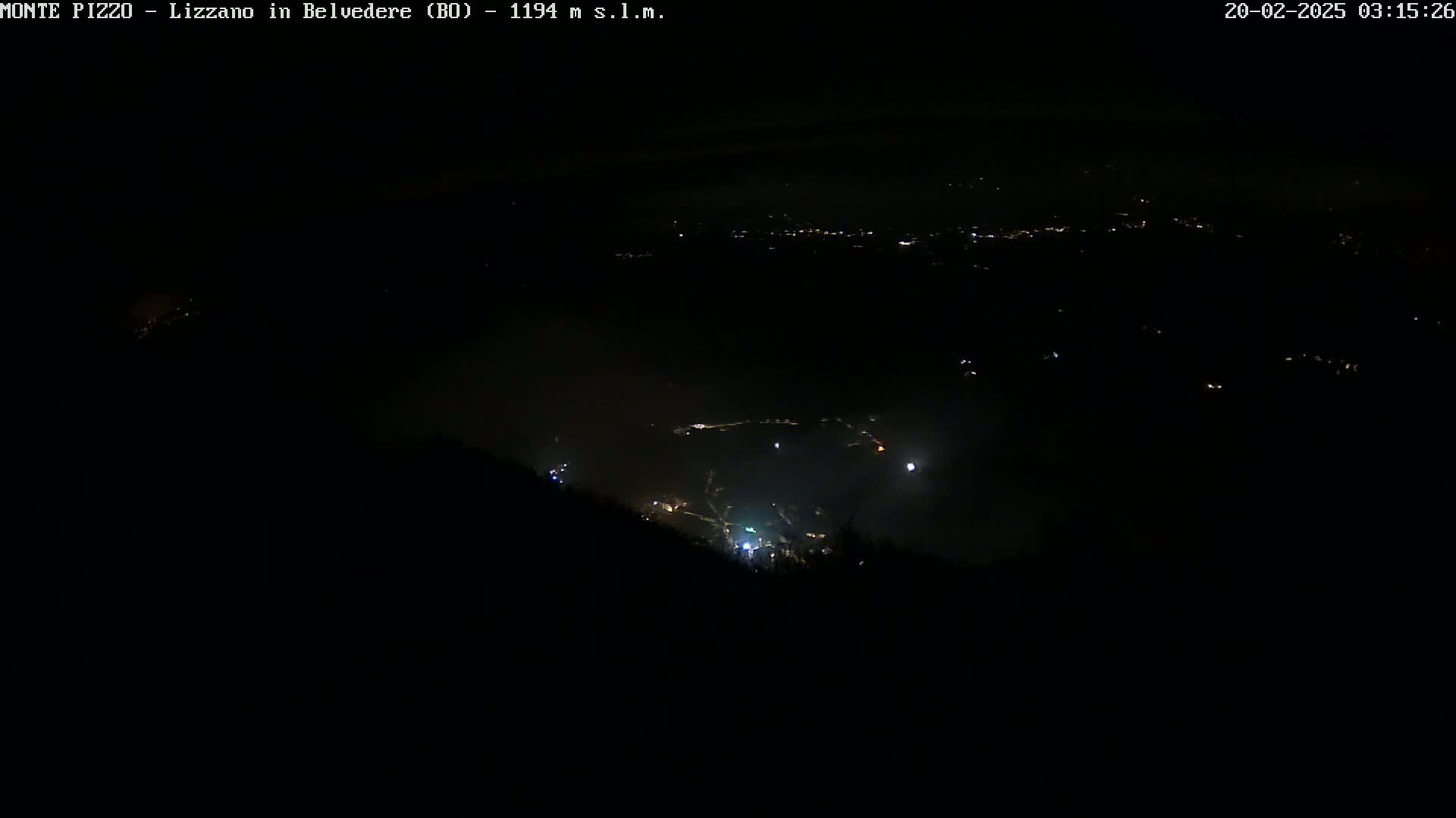 A nighttime, high-angle view shows a valley with scattered lights below a dark, foggy hillside.