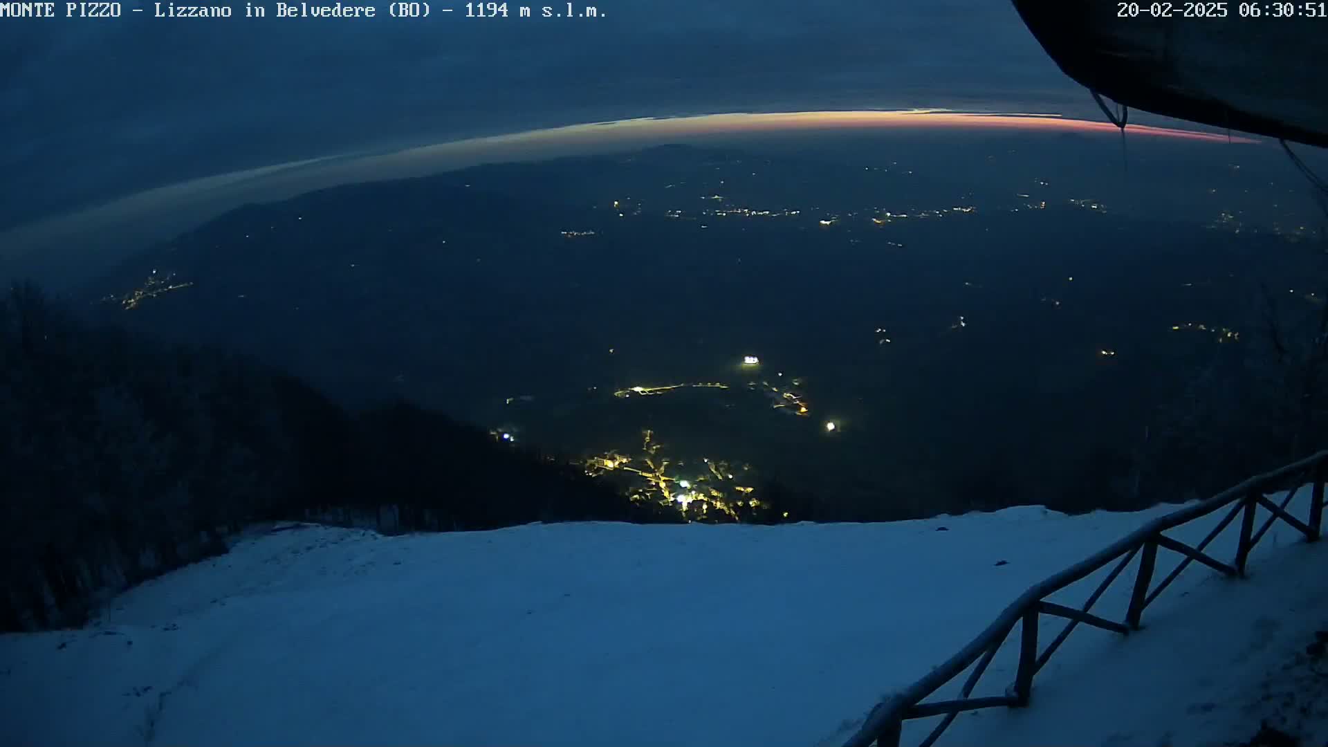 A nighttime, snowy mountaintop view shows a valley with scattered lights below, and a faint pinkish glow on the horizon.