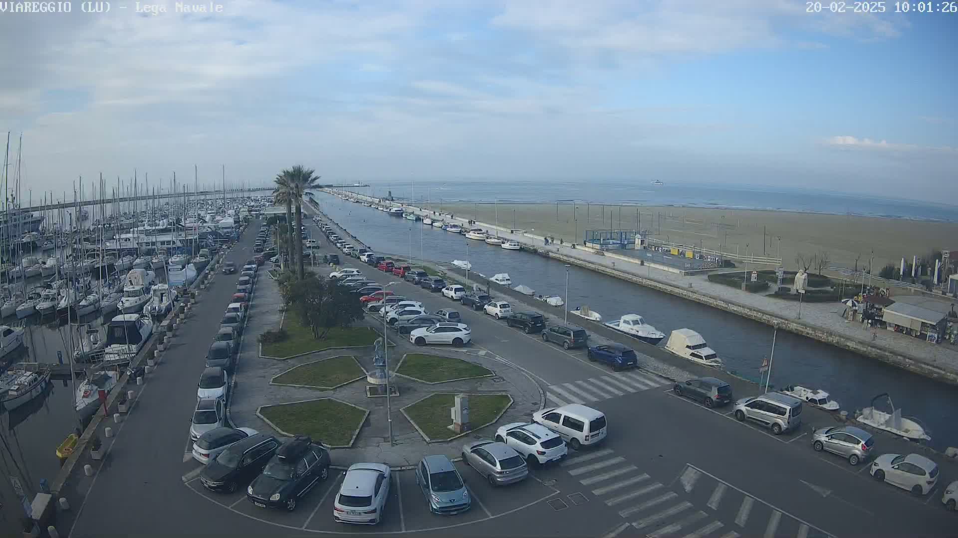 A marina filled with boats is next to a canal with parked cars along its edge, leading to a sandy beach under a partly cloudy sky.