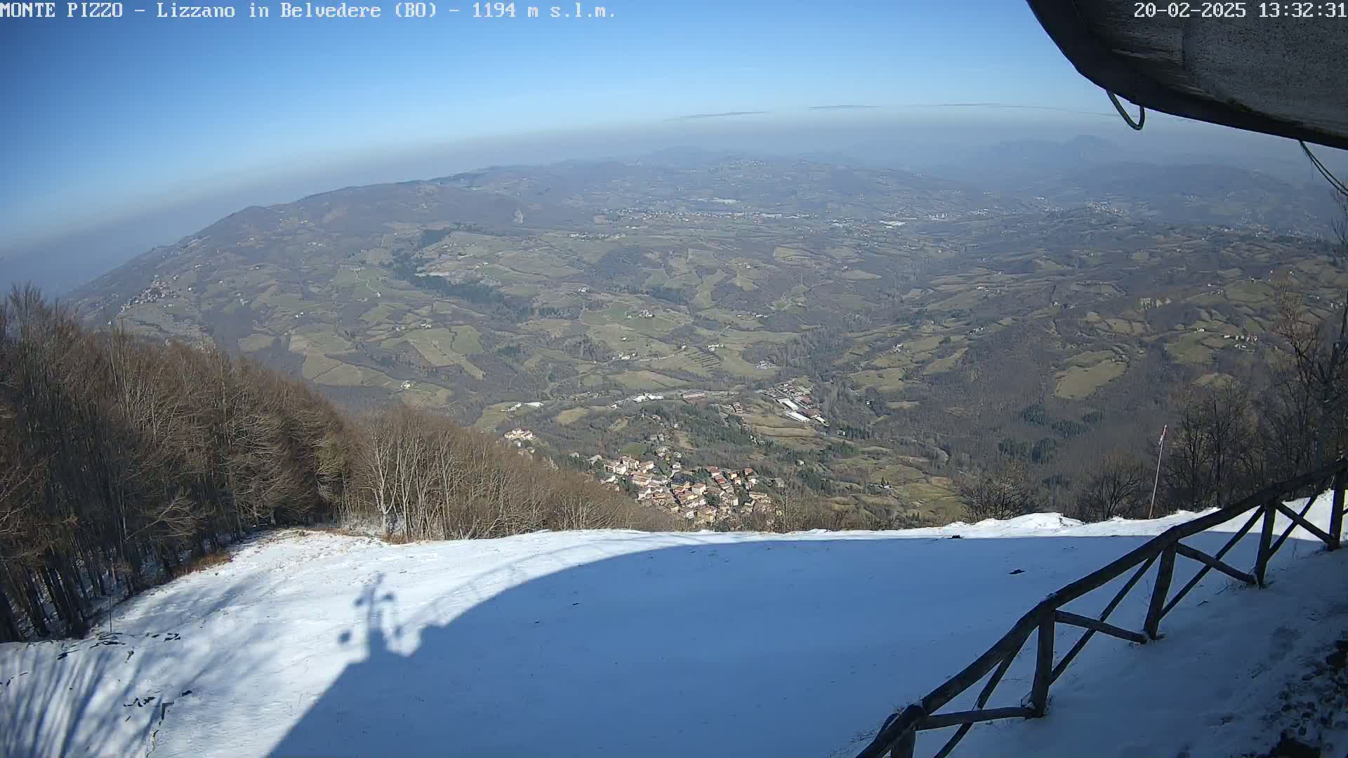 A snow-covered hillside overlooks a vast valley of farmland and a small village under a clear blue sky.