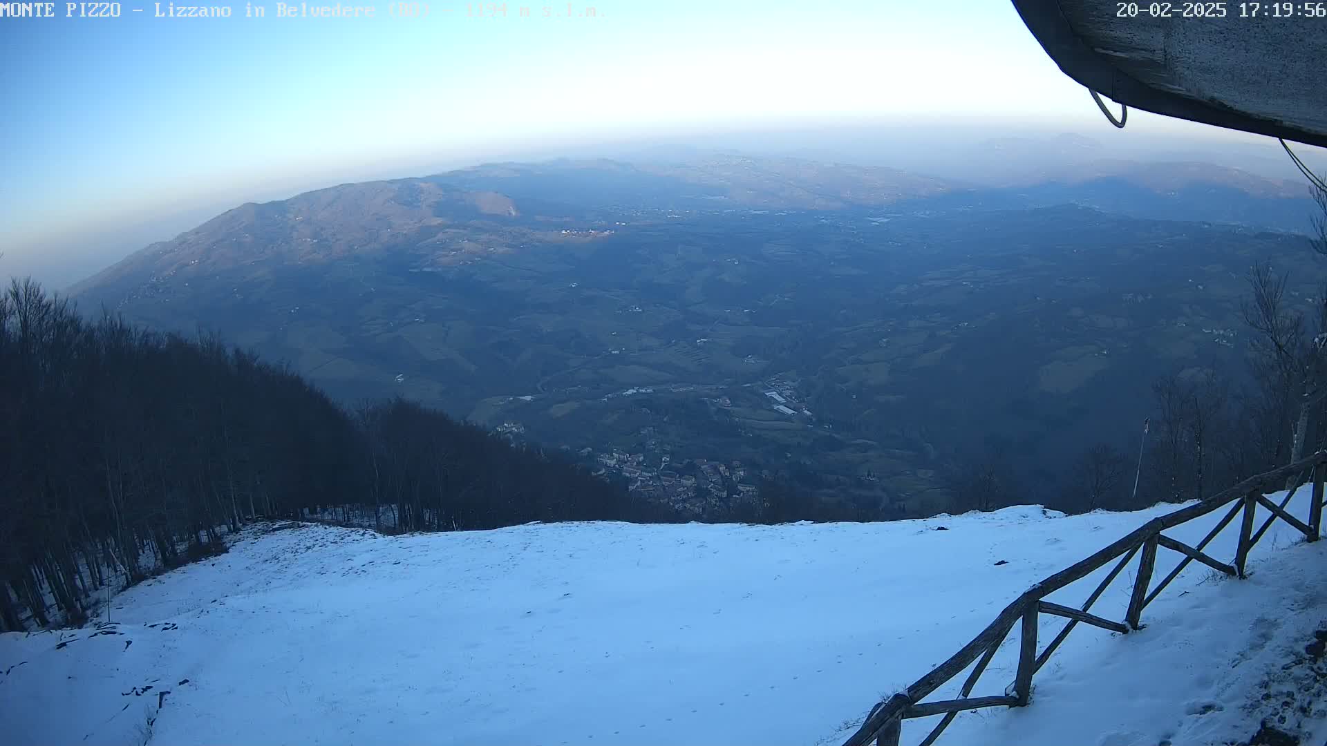 A snow-covered mountain slope overlooks a vast, hazy valley under a clear, pale sky.