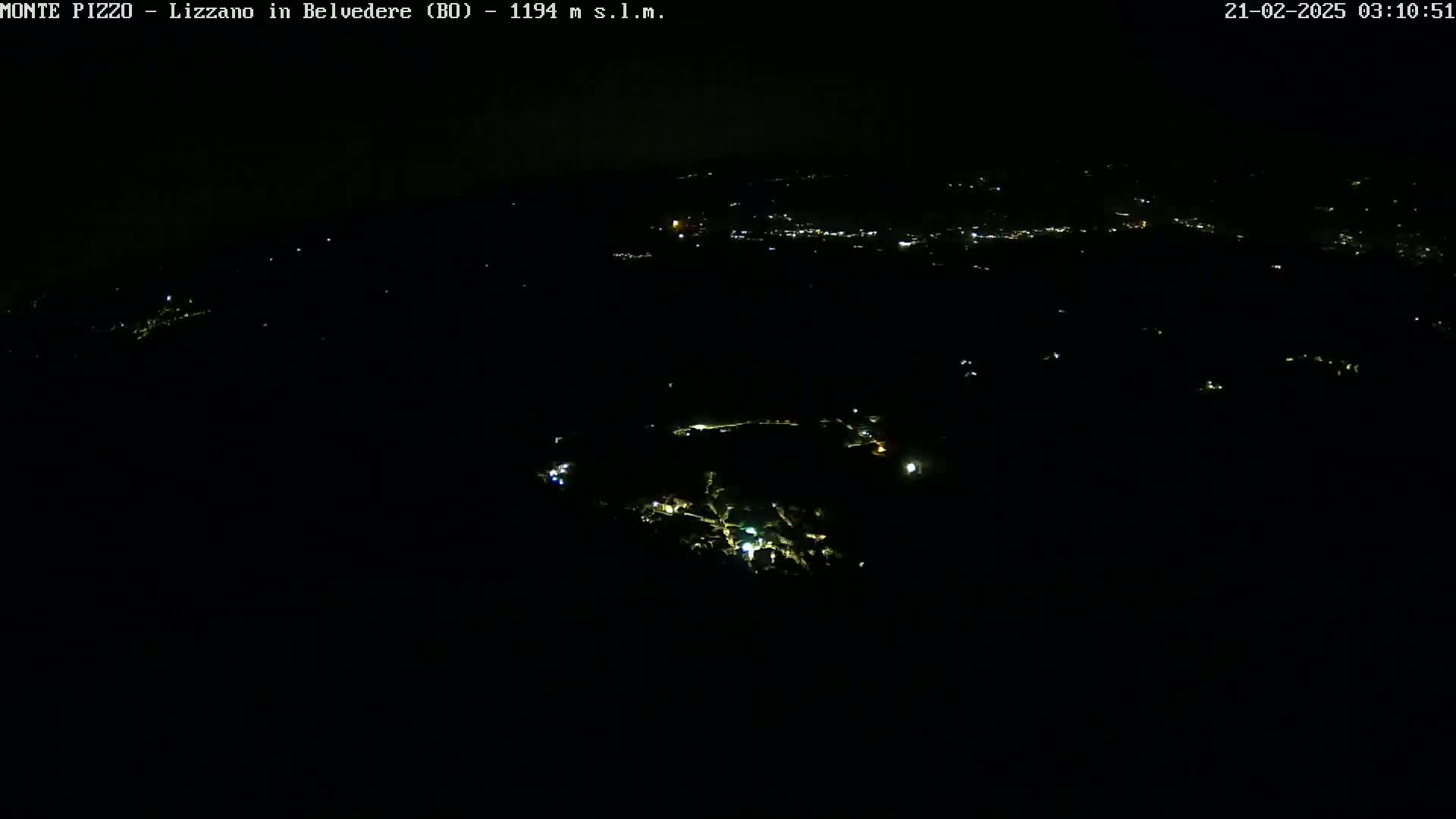 A nighttime, high-angle, long shot of sparsely lit, dark terrain with scattered points of light visible in the distance.