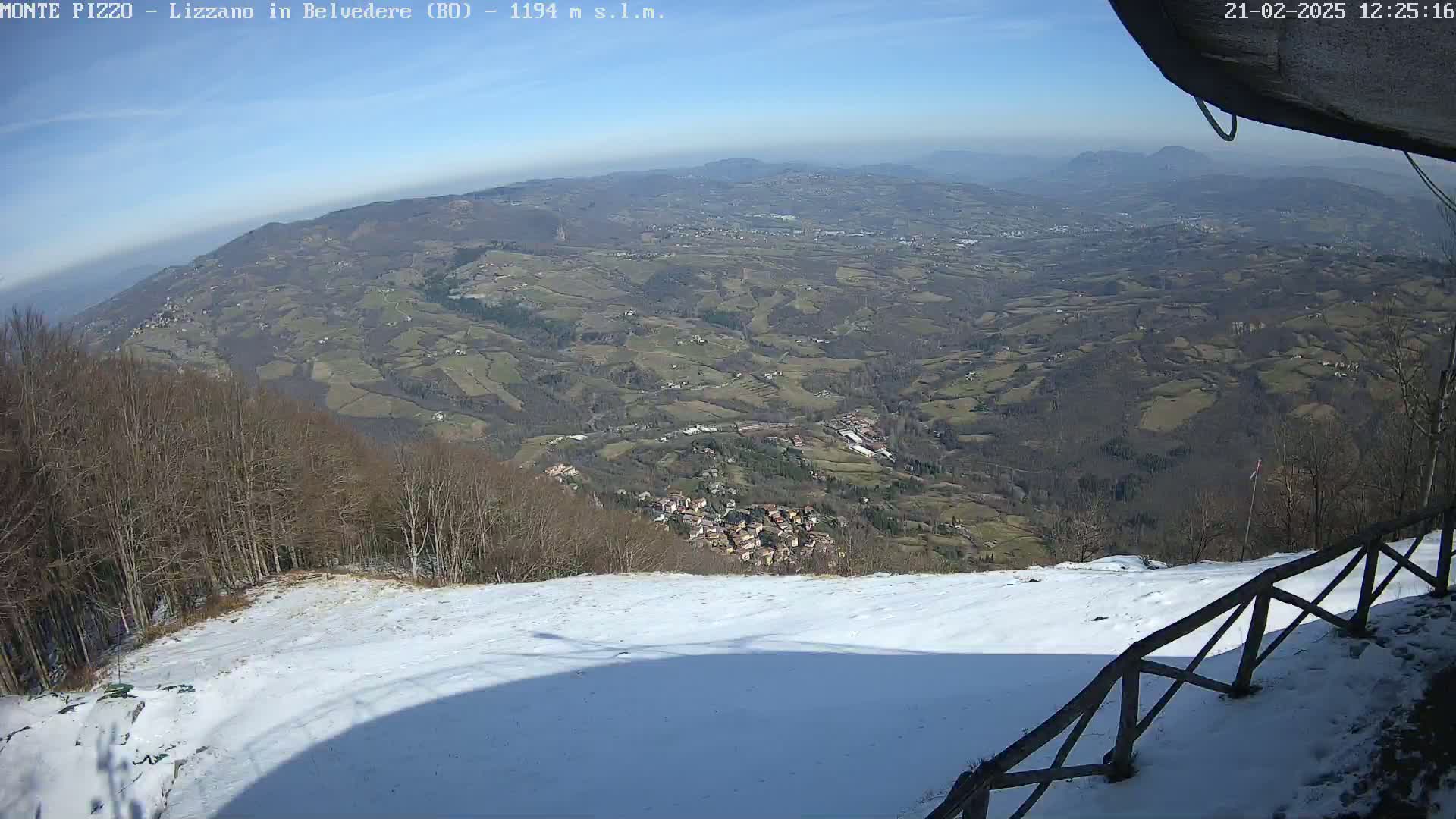 A snow-covered hillside overlooks a vast, hilly landscape under a clear, sunny sky.