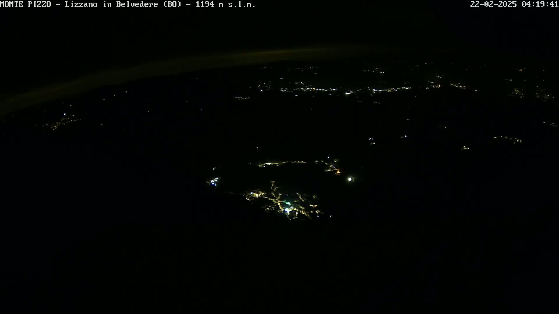 A nighttime, high-angle view shows a dark landscape with scattered points of light from buildings below.