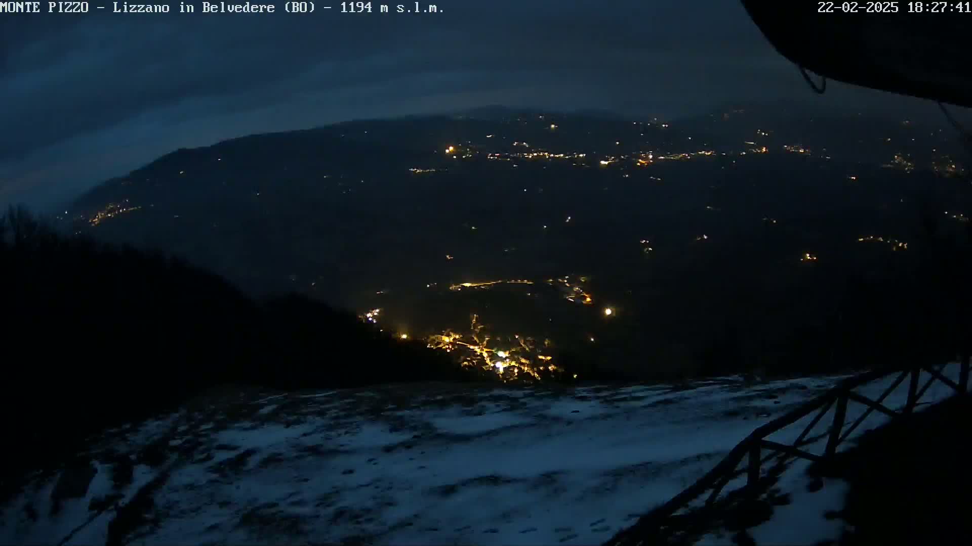 A nighttime view of a snow-covered landscape overlooking a valley with scattered lights from a town below under a dark sky.