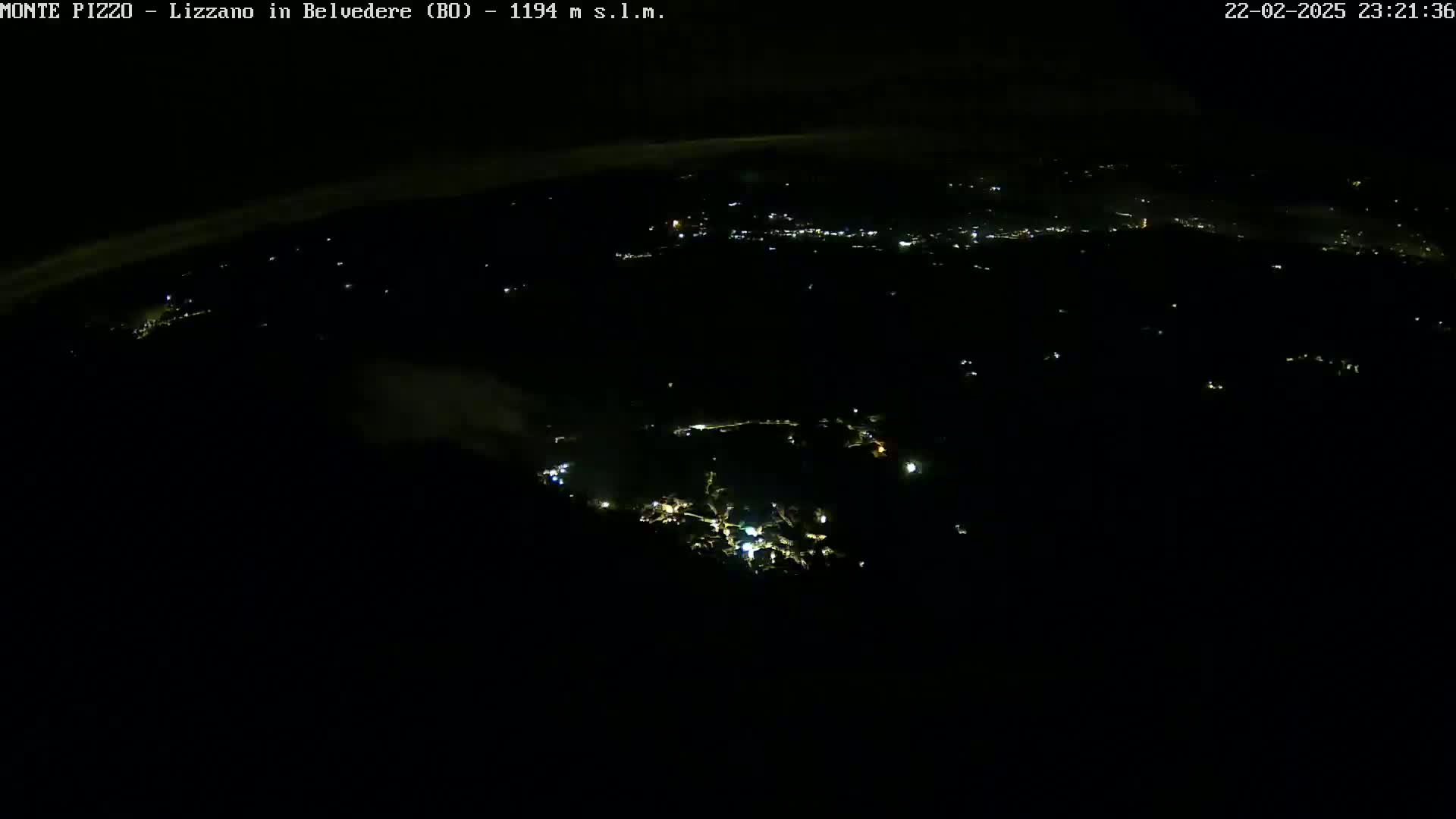 A nighttime, high-angle view shows a sparsely lit landscape with a small cluster of brighter lights in the center, possibly a town, under a dark, possibly cloudy sky.