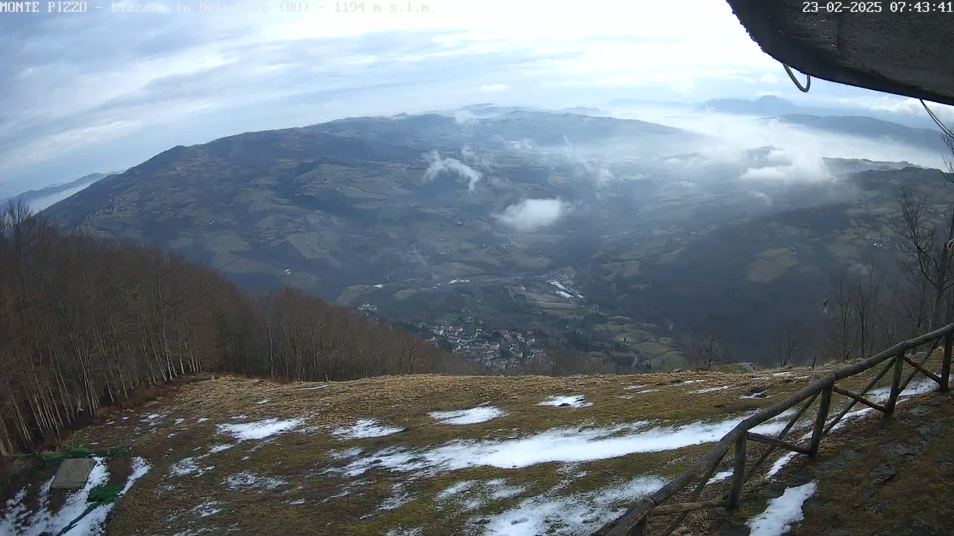 A high-angle view shows a valley shrouded in fog, with a small town visible below a snow-dusted hillside and a wooden fence in the foreground under a partly cloudy sky.