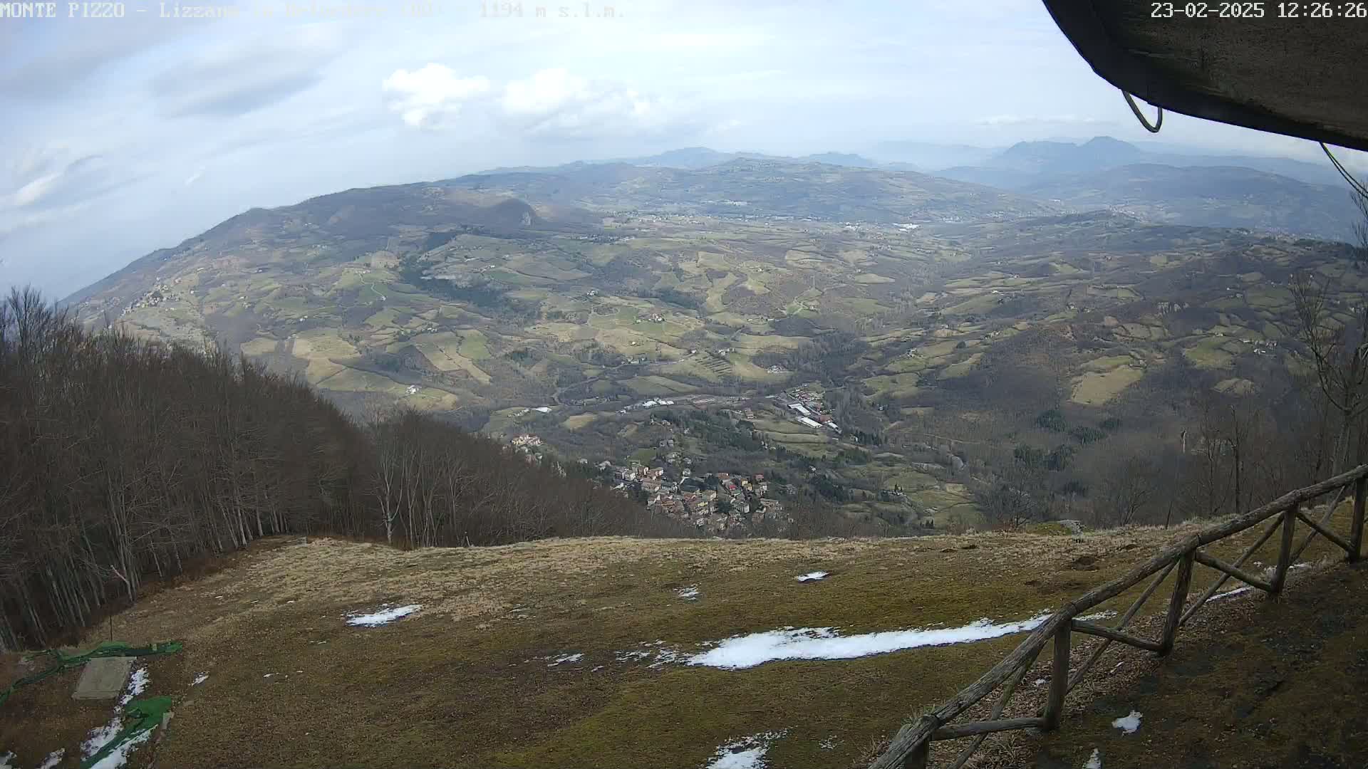 A high-angle view shows a sprawling valley of farmland and a small village under a partly cloudy sky, with patches of snow on the ground in the foreground.