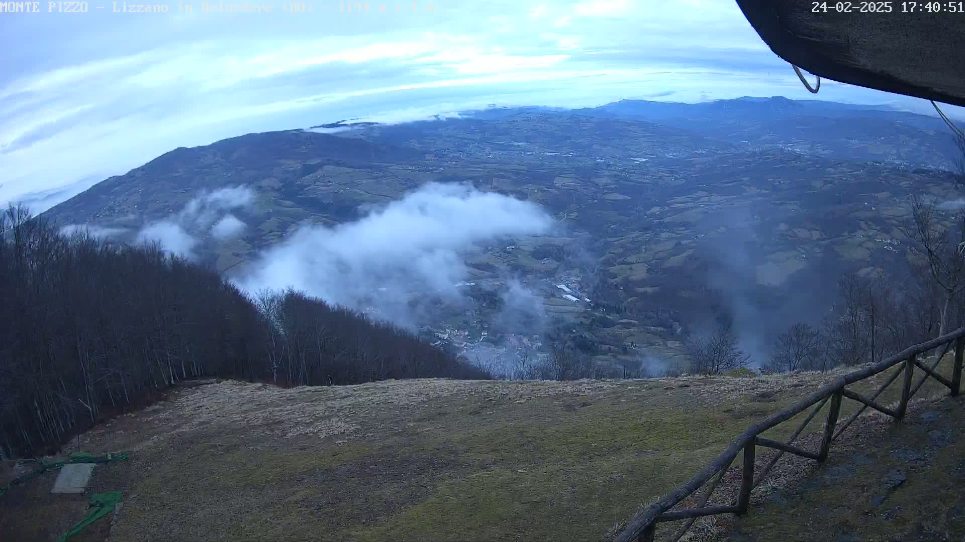 A high-angle view of a valley partially covered in fog, seen from a mountaintop with a wooden fence in the foreground under a mostly cloudy sky.