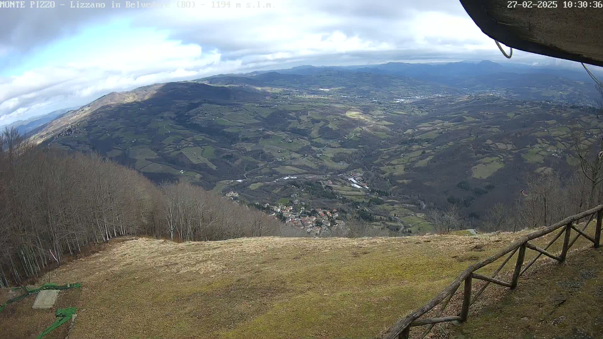 A high-angle view on a partly cloudy day shows a vast, rolling landscape of farmland and forest leading down to a small village, all observed from a high vantage point behind a wooden fence.