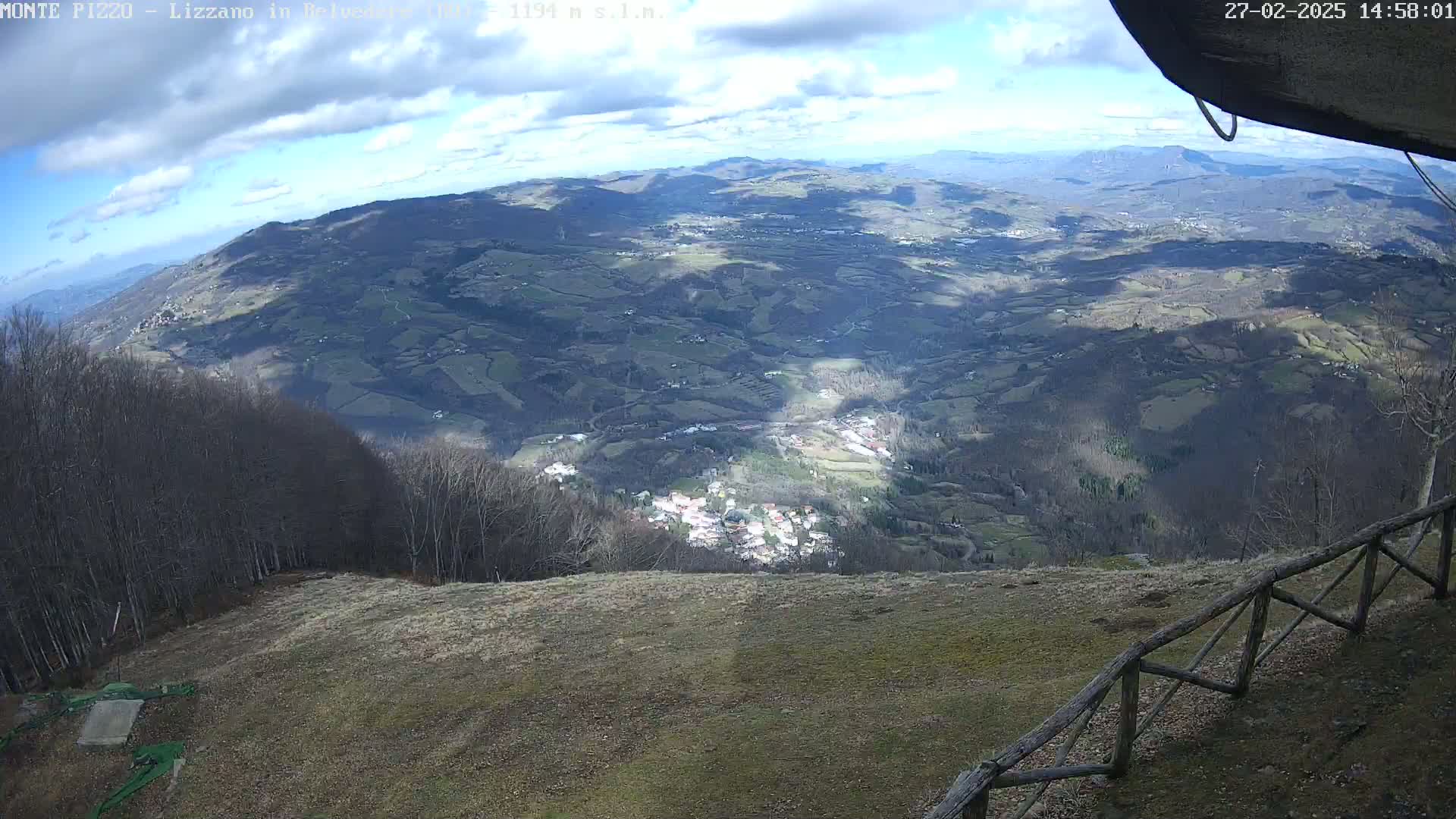 A high-angle view on a partly cloudy day shows a sprawling valley with a village nestled amongst rolling hills and forests, all seen from a mountaintop vantage point with a wooden fence in the foreground.