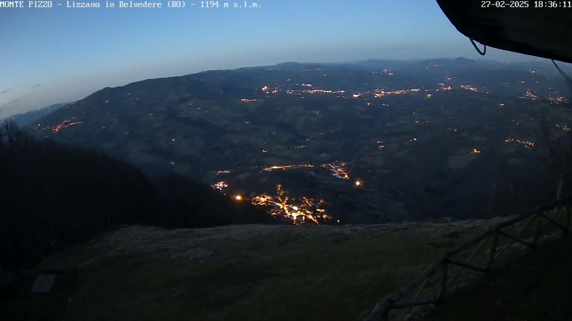 A nighttime, high-angle view of a valley with scattered lights from a town below, under a twilight sky.