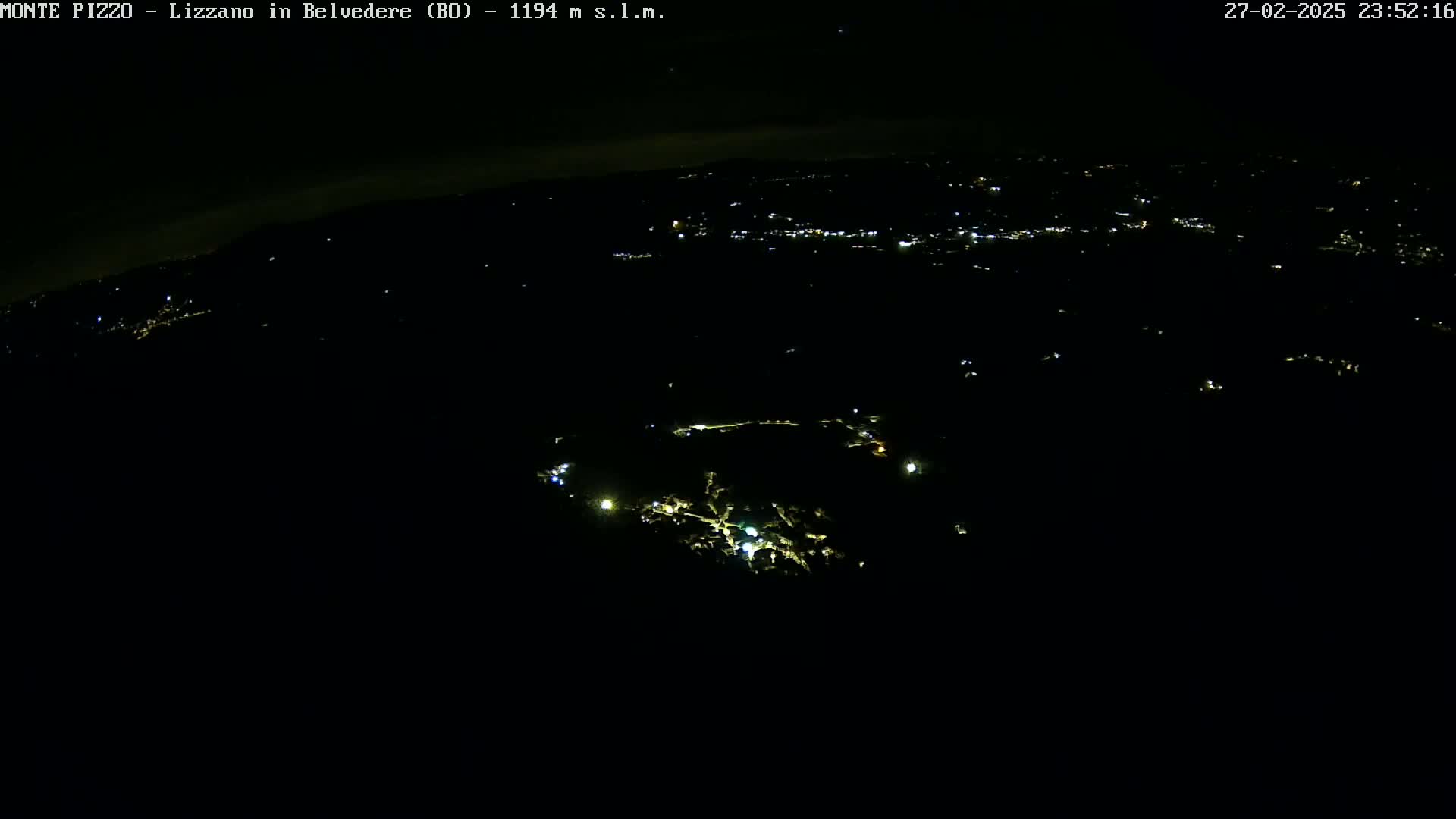 A nighttime, aerial view shows a dark landscape with scattered lights from a town in the distance and a few closer lights, possibly from buildings or roads.