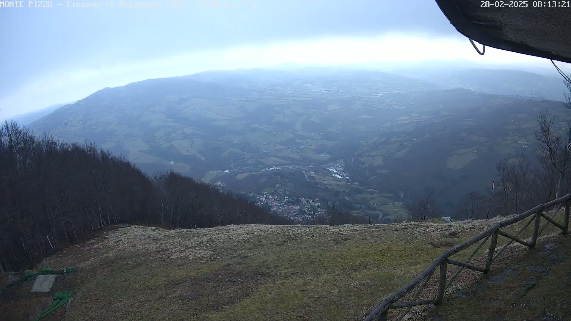 A high-angle, long shot of a mountainous landscape under an overcast sky shows a distant village nestled in a valley below a high vantage point with a wooden fence in the foreground.