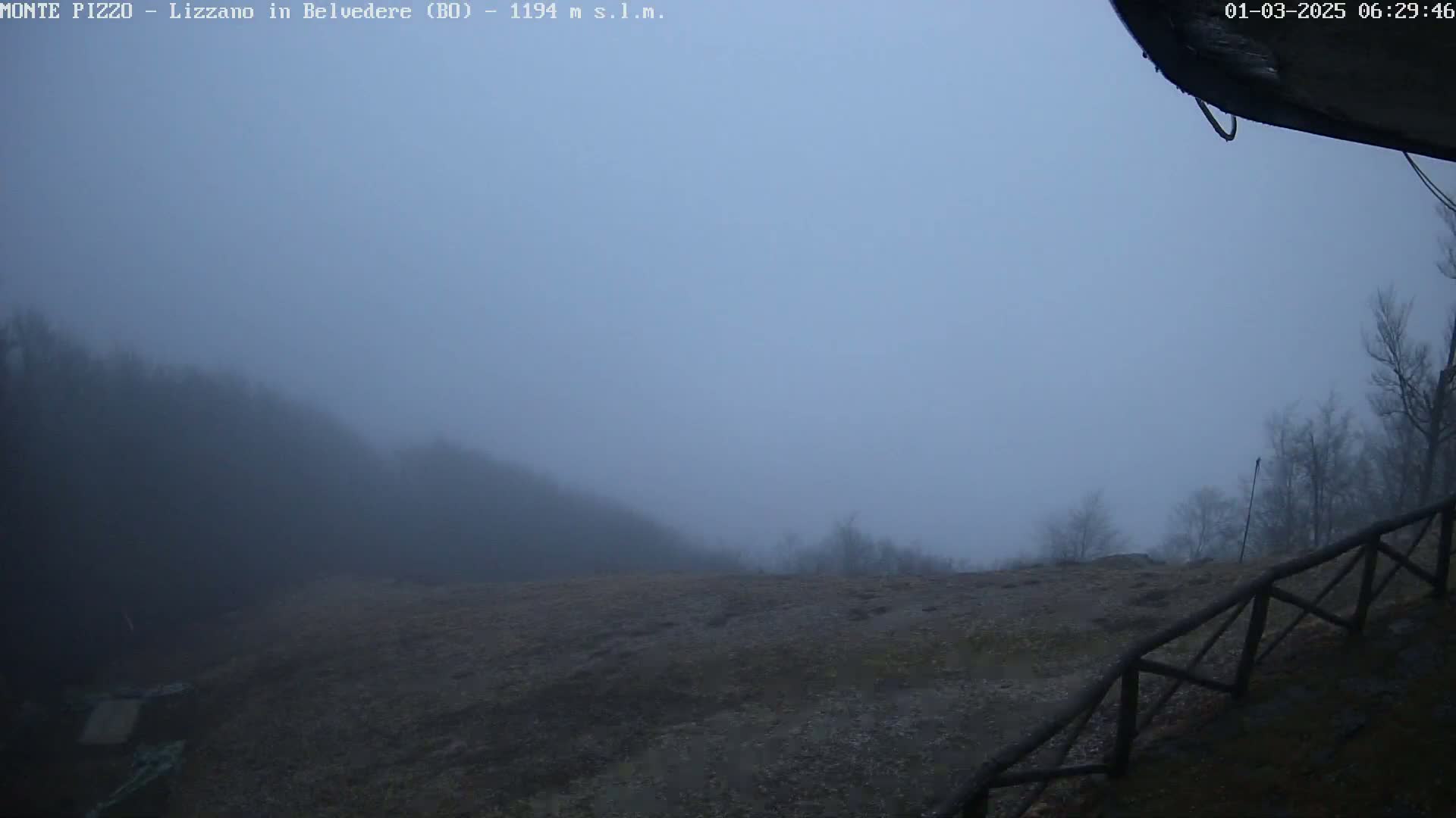 A foggy, gray landscape with a rocky foreground and a low-lying hill shrouded in mist in the background, partially obscured by a wooden fence.