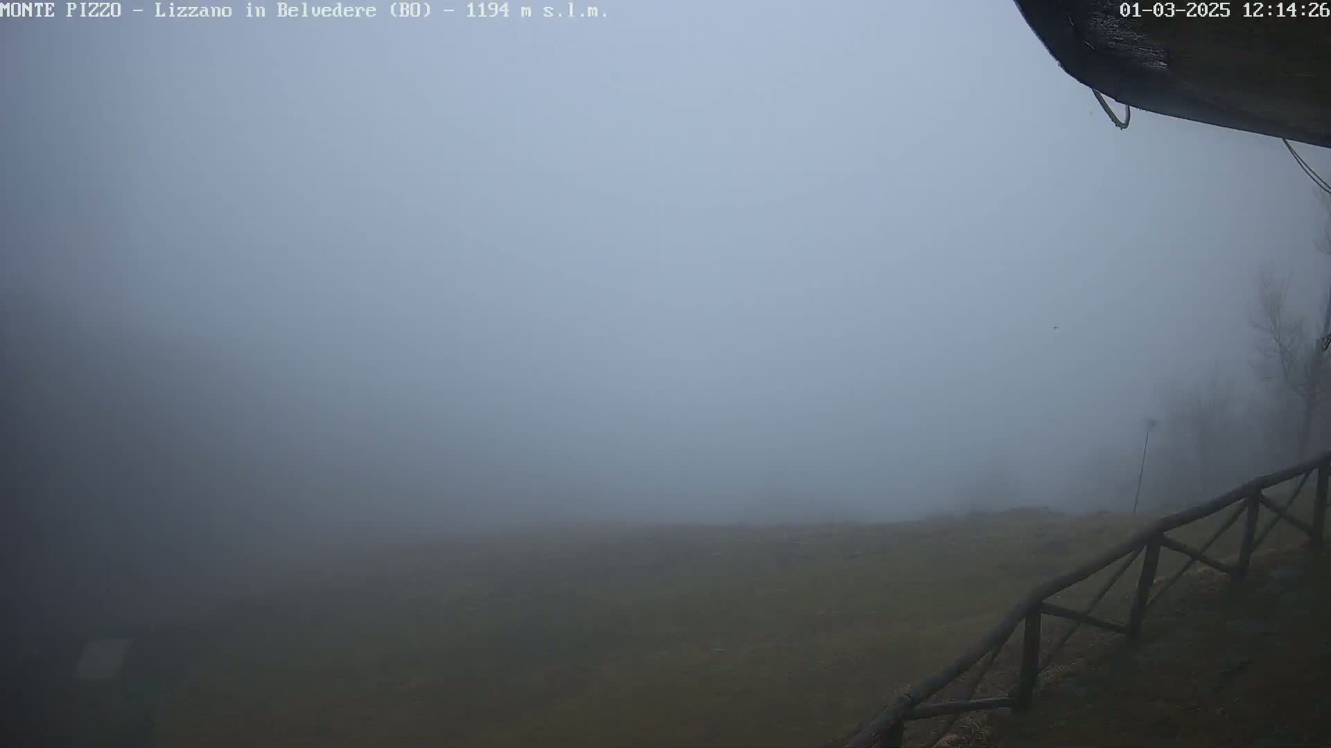 A foggy, outdoor scene shows a dark, flat expanse of land partially obscured by heavy fog, with a wooden fence in the foreground and a small portion of a building visible in the upper right corner.
