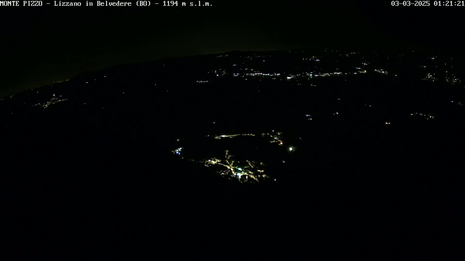A nighttime aerial view shows a dark landscape with scattered lights from a town or village in the distance.