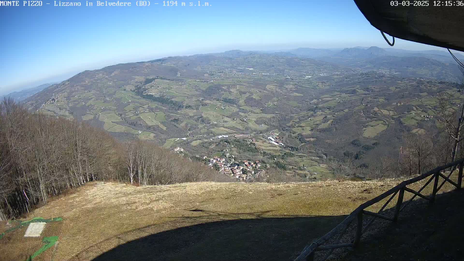 A high-angle, long shot on a sunny day shows a sprawling valley of farmland and a small village, viewed from a high vantage point with a wooden fence in the foreground.