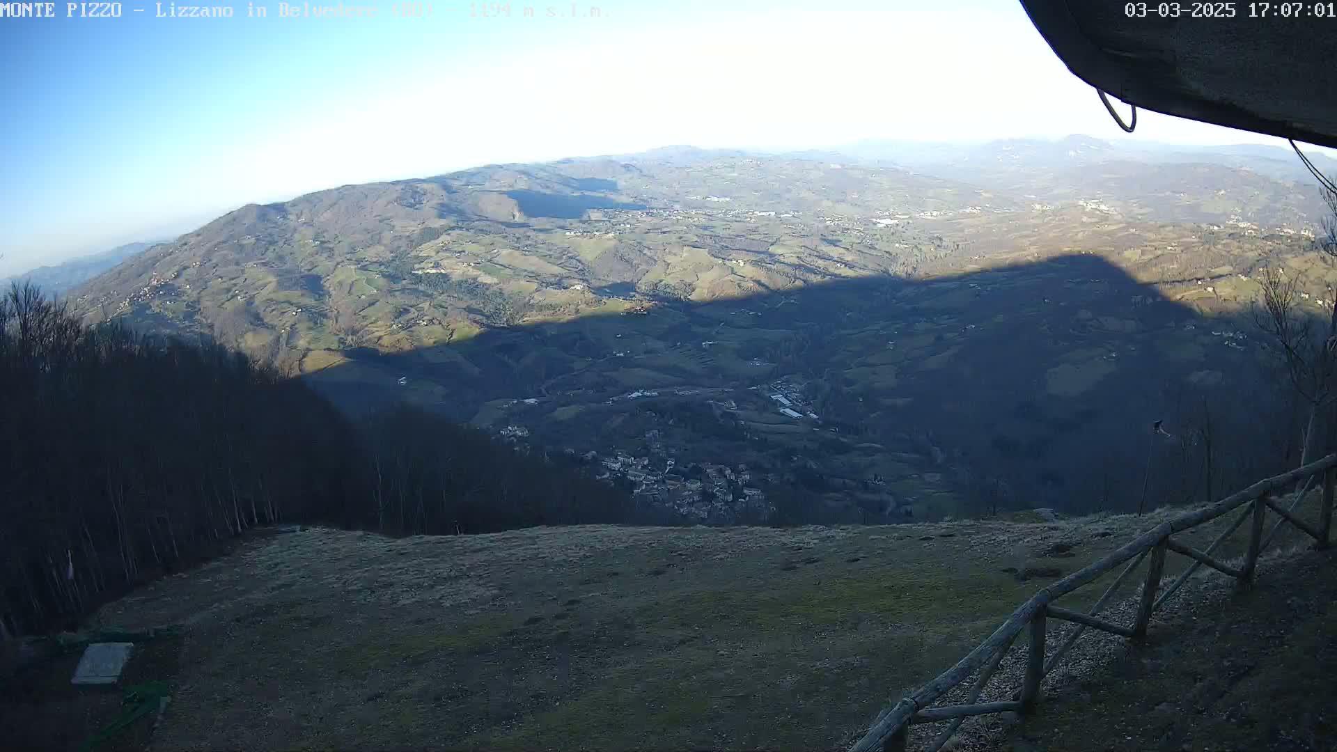 A high-angle, long shot on a clear, sunny day shows a vast, hilly landscape with a village nestled in the valley below, partially cast in shadow.