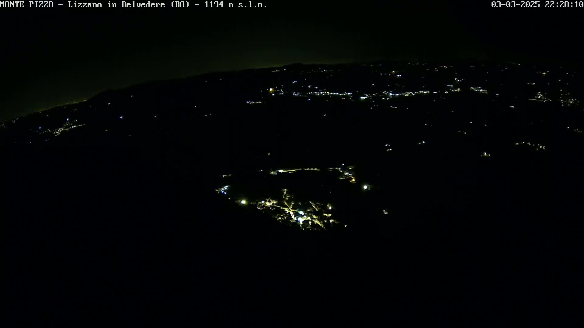 A nighttime, high-angle view shows a dark landscape with sparsely scattered lights from a town or village below.
