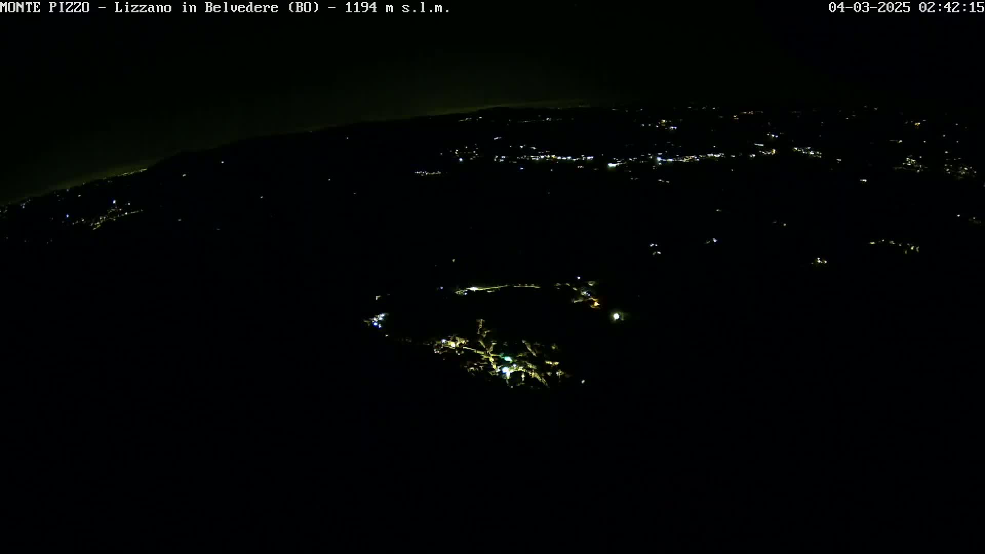 A nighttime, high-angle view shows a dark landscape with scattered lights from a town or village in the distance.