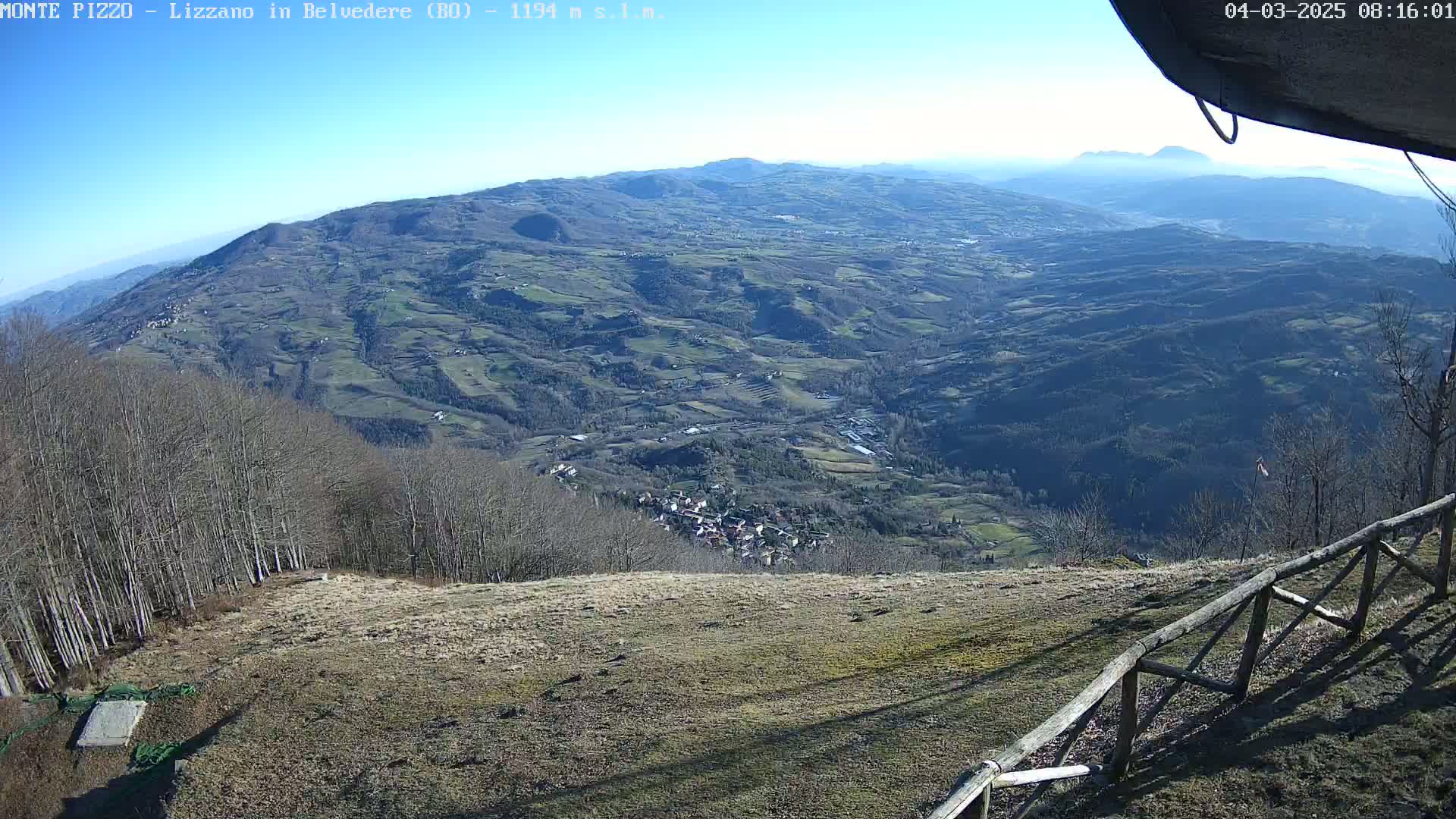 A high-angle, long shot on a clear, sunny day shows a vast, hilly landscape with a small town nestled in a valley, partially obscured by leafless trees in the foreground.