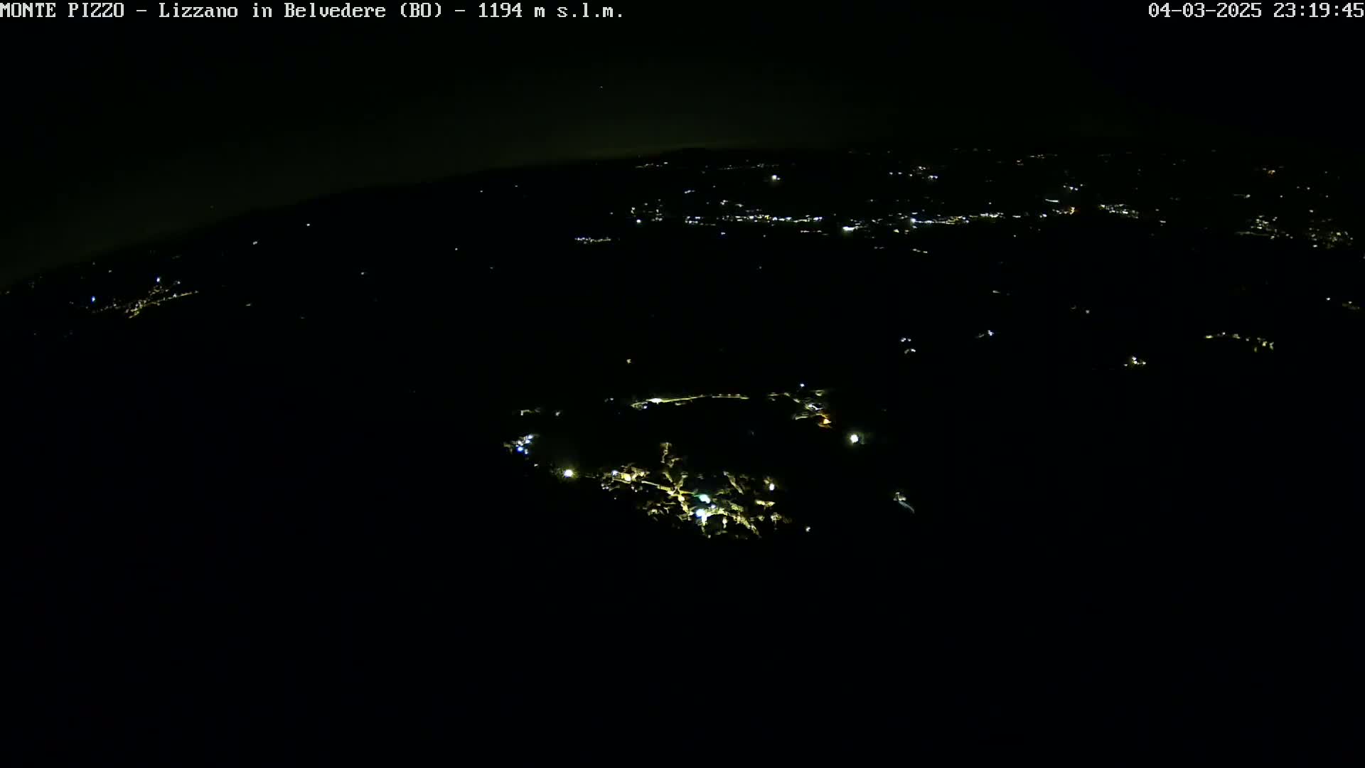 A nighttime, high-angle view shows a sparsely lit landscape with a few clusters of lights below, possibly a town or village, under a dark sky.